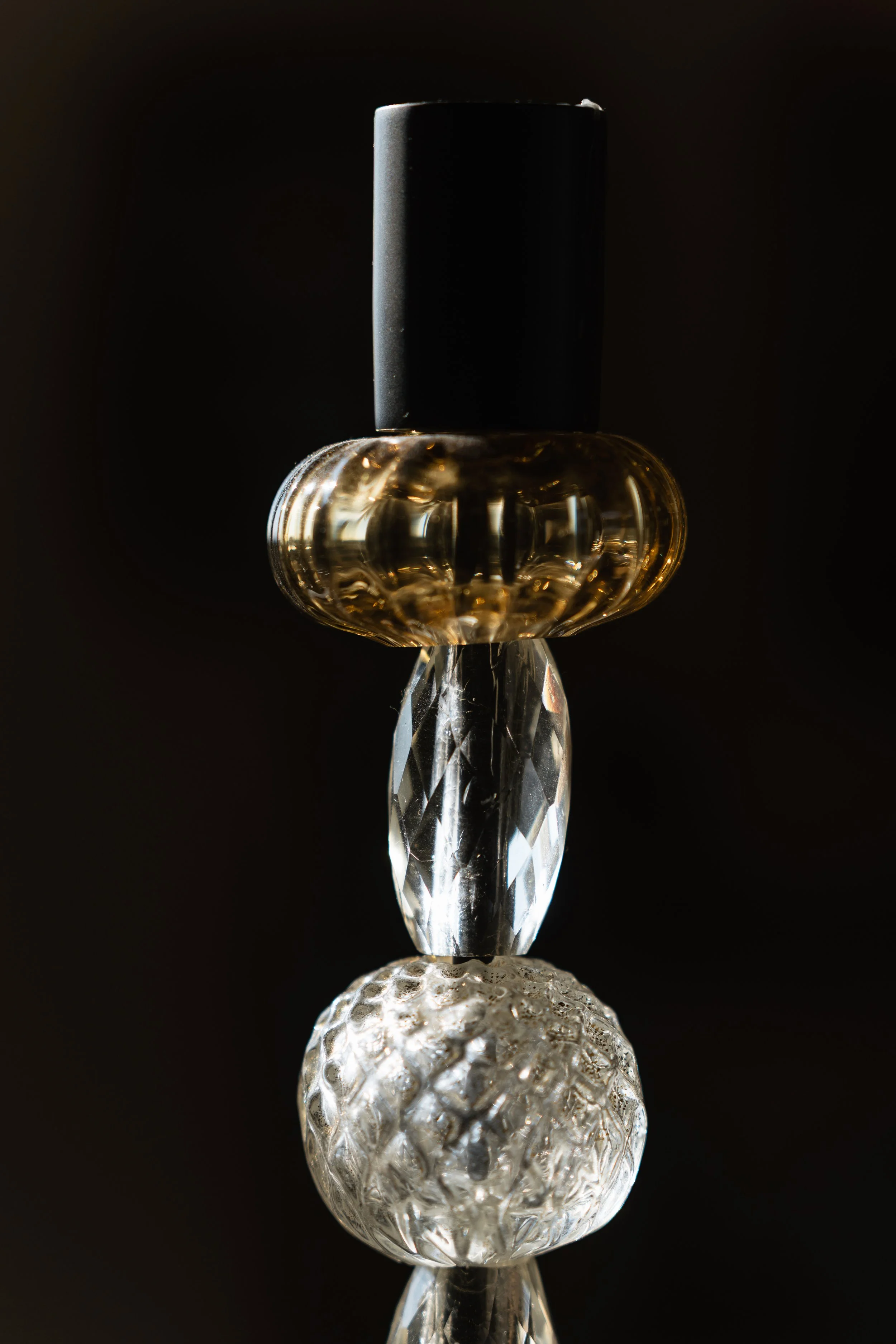 Close-up of glass decorative objects arranged in a vertical line against a dark background, including a textured, frosted glass ball at the bottom, a faceted clear glass piece, a small reflective glass object, a round glass ring, and a black cylindri