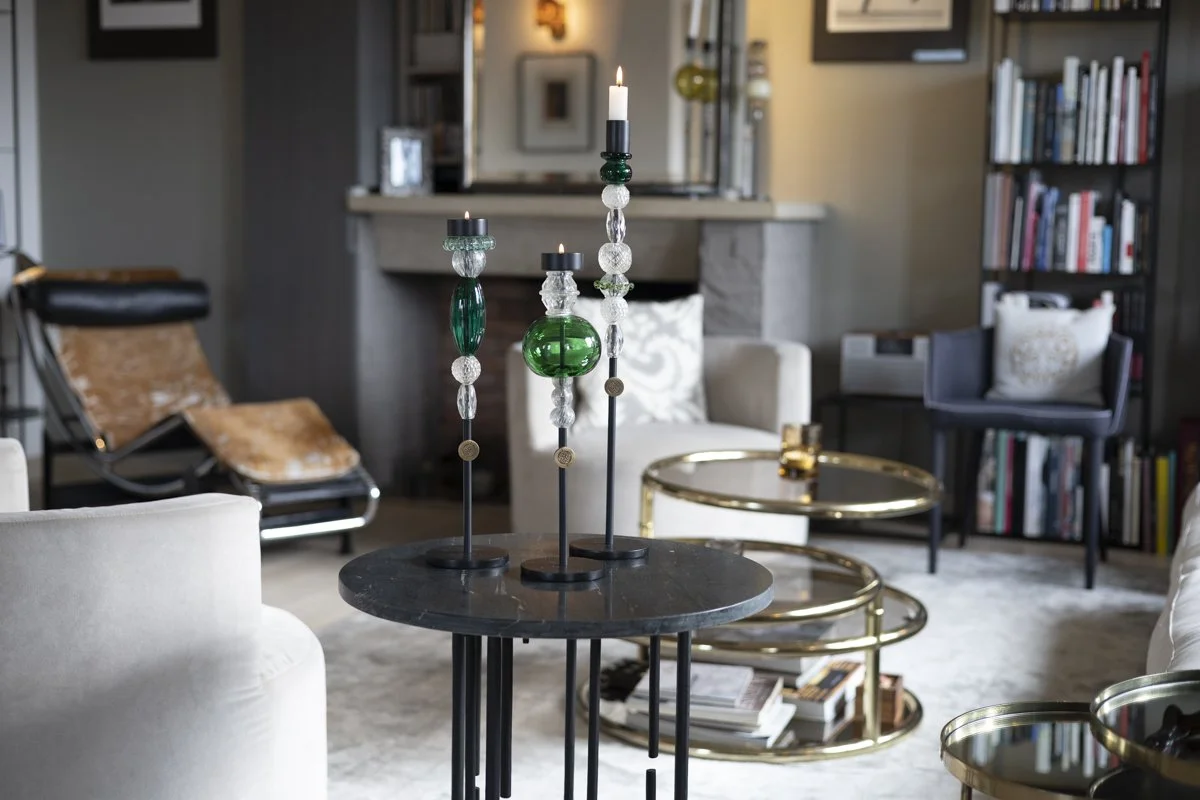 Living room with a black marble side table holding three tall candle holders with candles, surrounded by white and dark furniture, gold nesting tables, a bookshelf, and a light-colored rug.