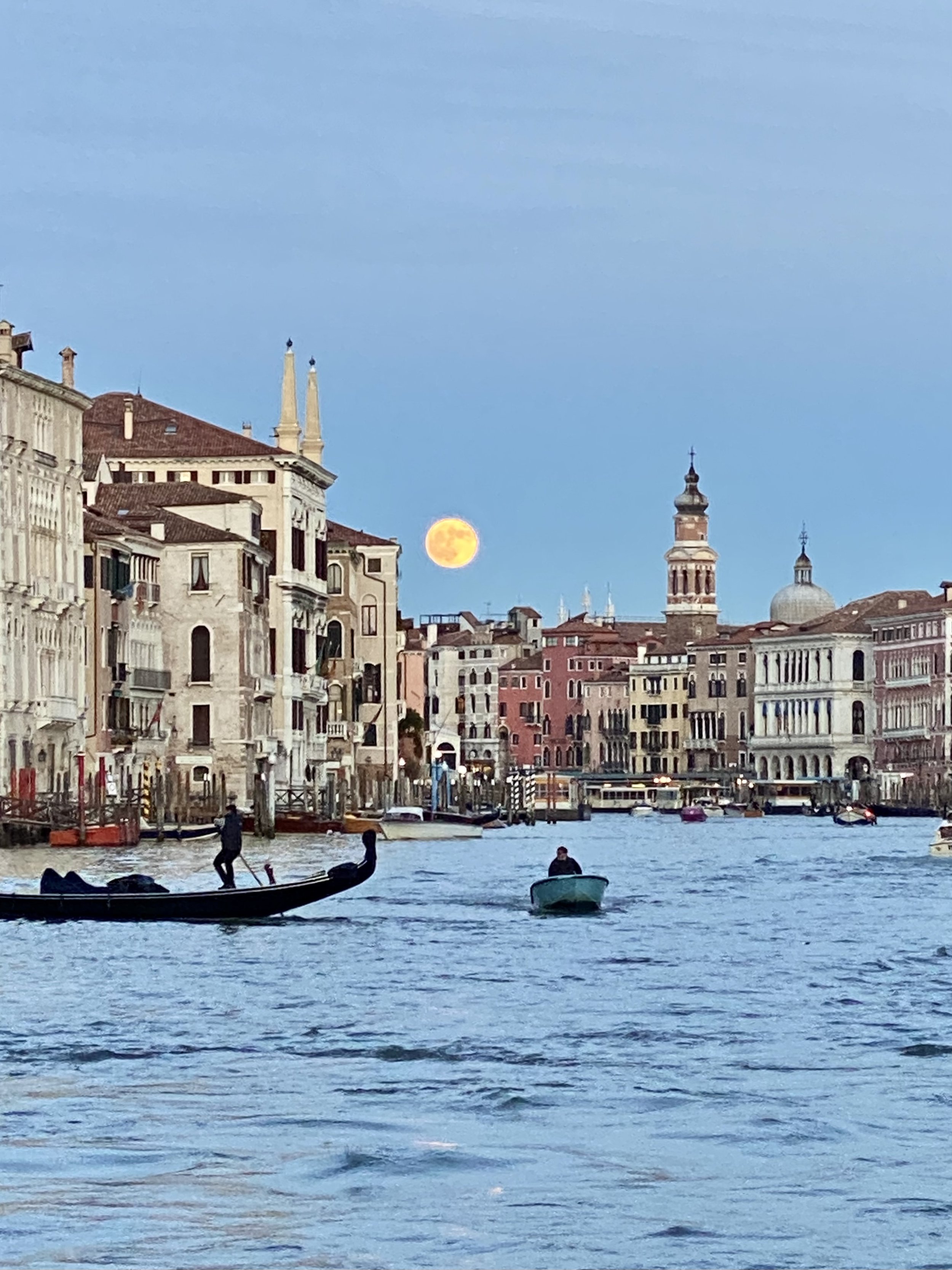 A view of the Grand Canal in Venice, Italy, with gondolas on the water and historic buildings along the shoreline, under a daytime sky with the moon visible in the background.