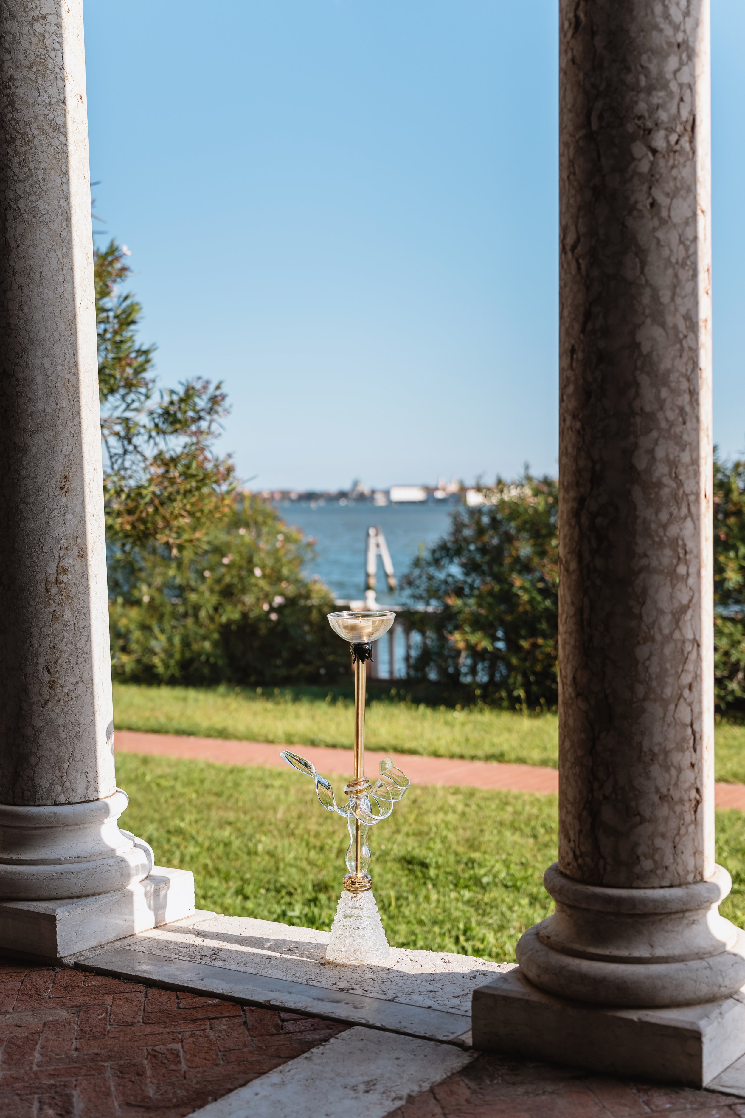 View of a waterfront through a stone and columned structure, with an ornate glass candle holder in the foreground.