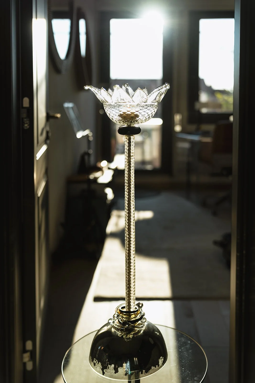 A decorative glass object resembling a flower, mounted on a tall, textured stand with a black base, placed on a glass table in front of a window with sunlight streaming in.