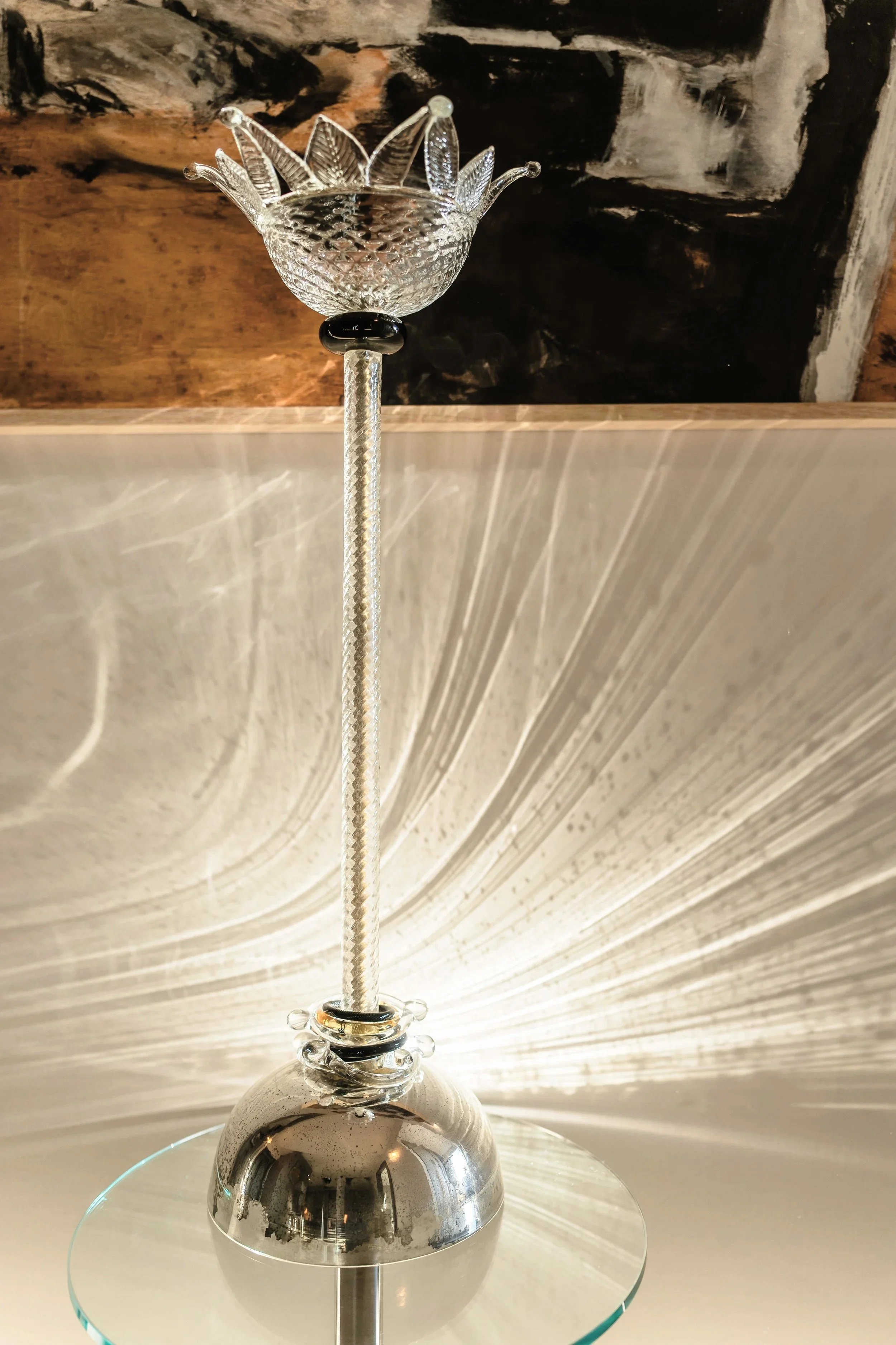 A decorative glass sculpture with a textured bowl and leaf-shaped extensions, supported by a textured stem and a metallic base, placed on a round glass surface with a textured light-colored background.