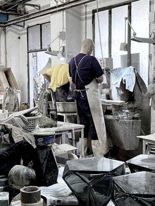 Two workers in a cluttered industrial kitchen or workshop, one using a large machine with cloth draped over it, surrounded by various supplies and equipment.