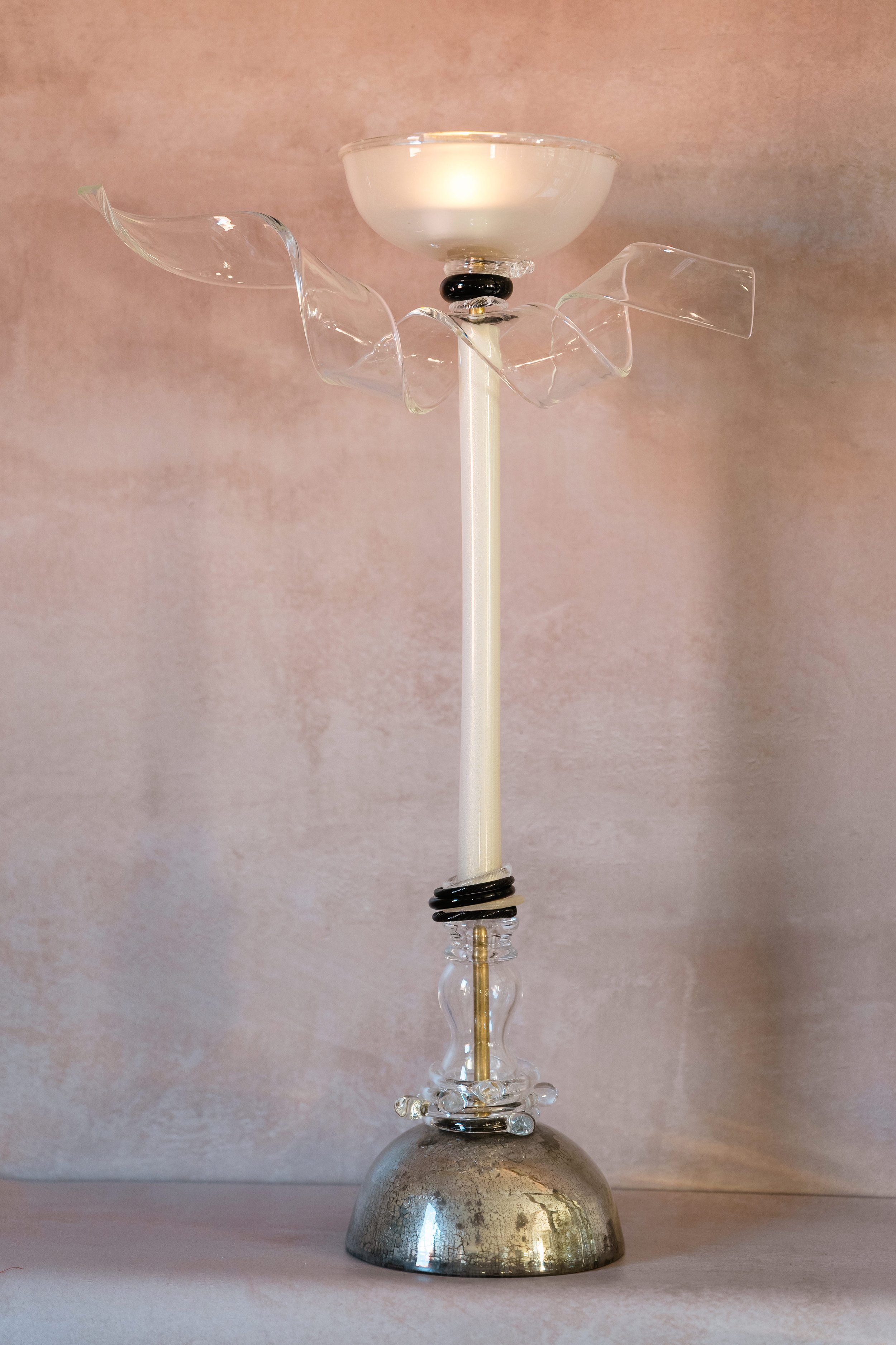A decorative candle holder with a lit white candle on a backdrop of light pinkish-beige wall.