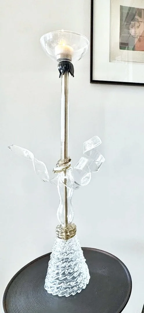 Glass lamp with a lit candle on top, featuring a sculptural base and abstract glass design, placed on a black round surface.
