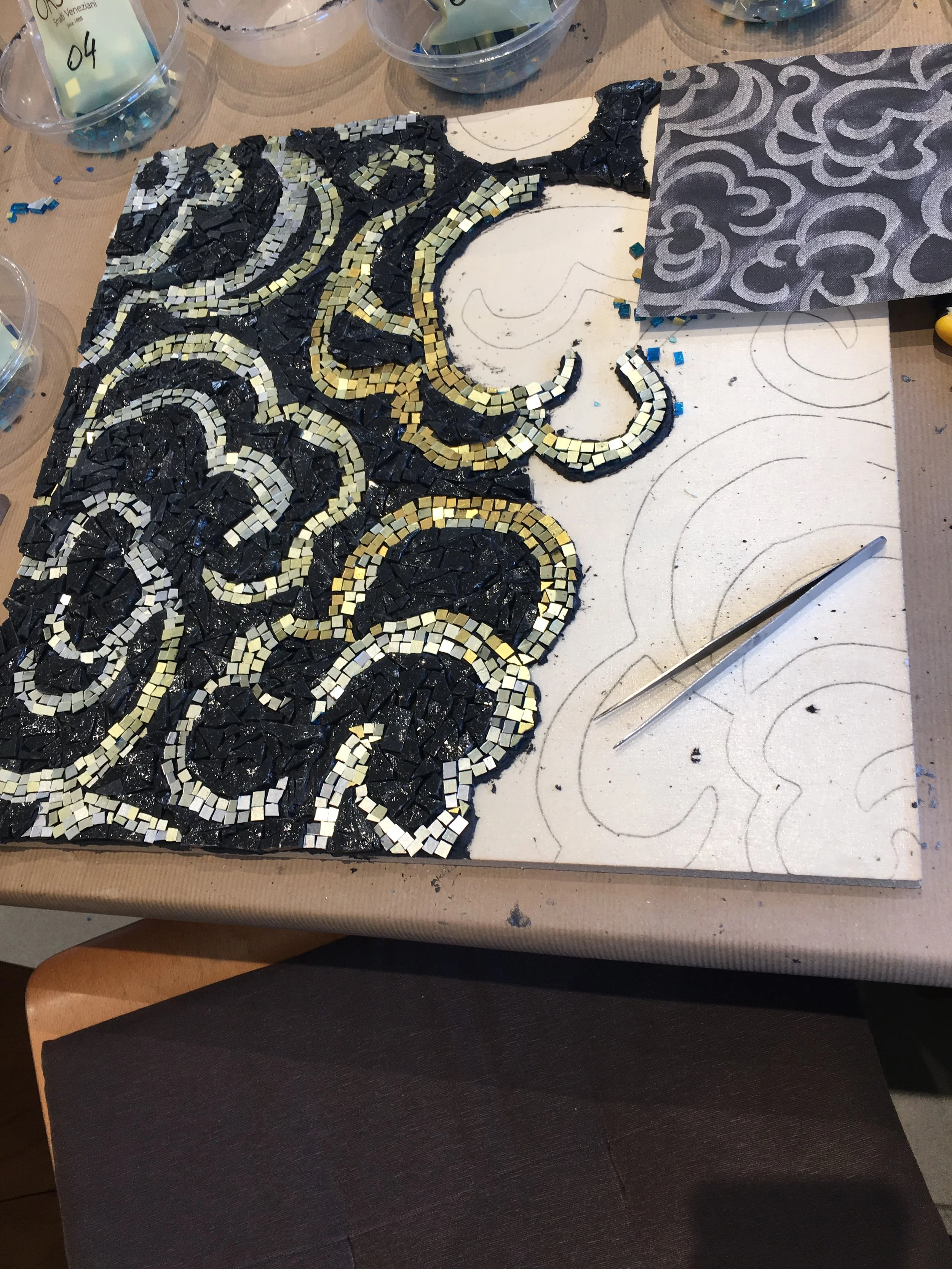 A mosaic design in progress made with small square tiles, with some black, white, and gold tiles on a white backing board. The design features swirling patterns and curves, with some sketches and patterns drawn on the board. There are small bowls and containers with mosaic tiles around the workspace.