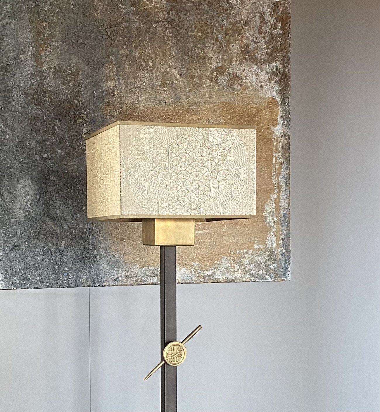 A modern floor lamp with a rectangular textured lampshade, featuring intricate patterns, against a textured wall and a white wall background.