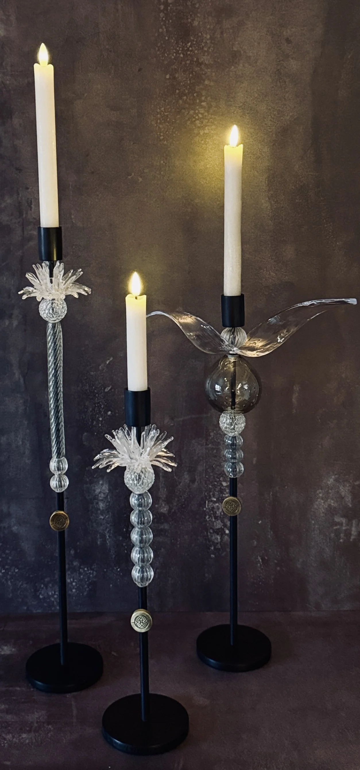 Three tall glass and metal candlesticks with white candles, some lit, on a dark surface against a dark textured background.