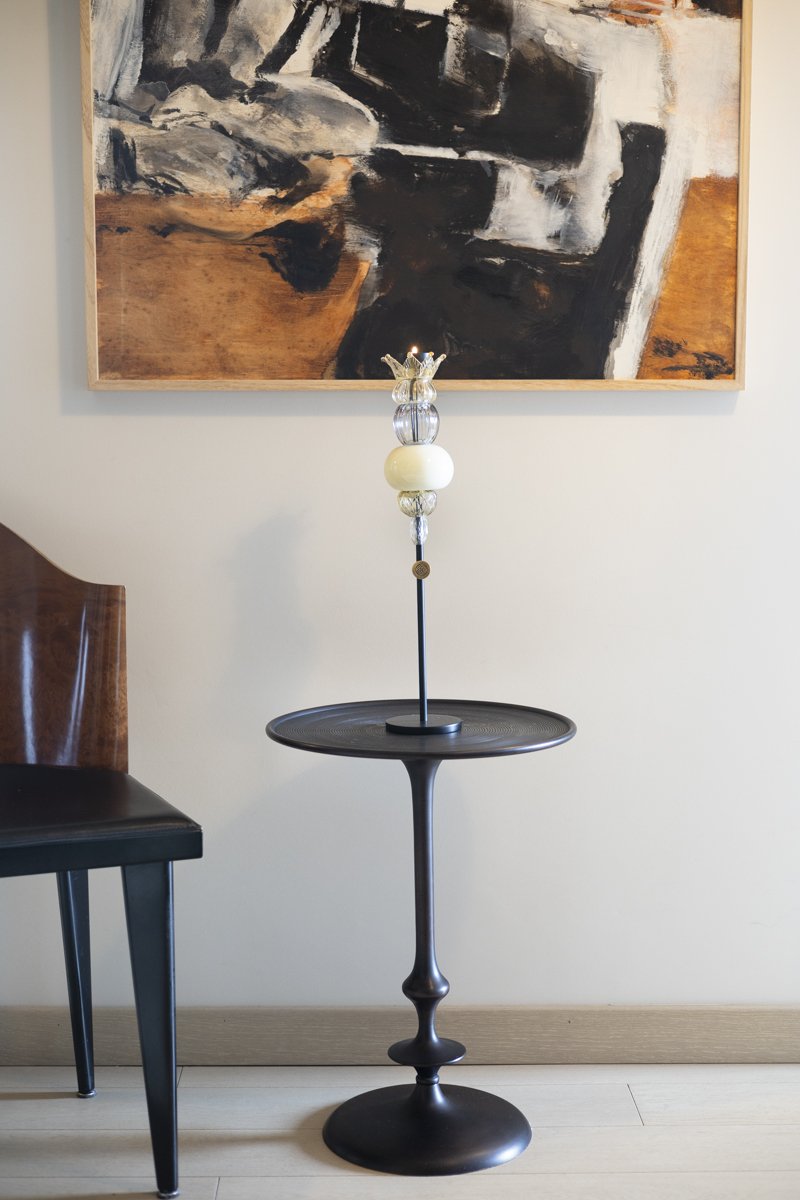 A modern, black metal side table with a decorative glass lamp on top, positioned next to a wooden chair, with an abstract painting with black, white, gray, and brown tones hanging on a white wall behind.