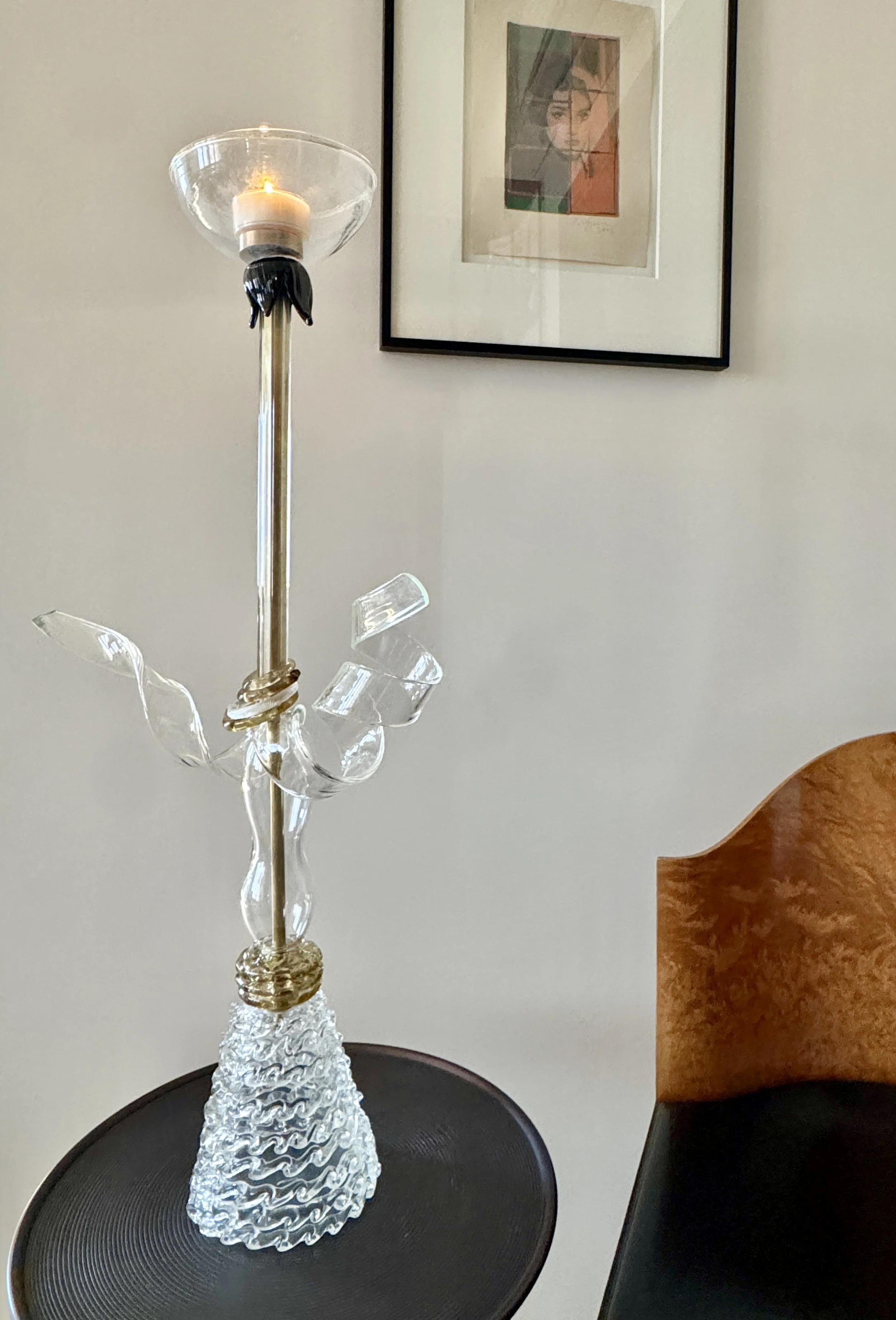 A glass and crystal table lamp with a candle inside on a round black table next to a wooden chair, with a framed artwork of a woman's face hanging on the wall in the background.