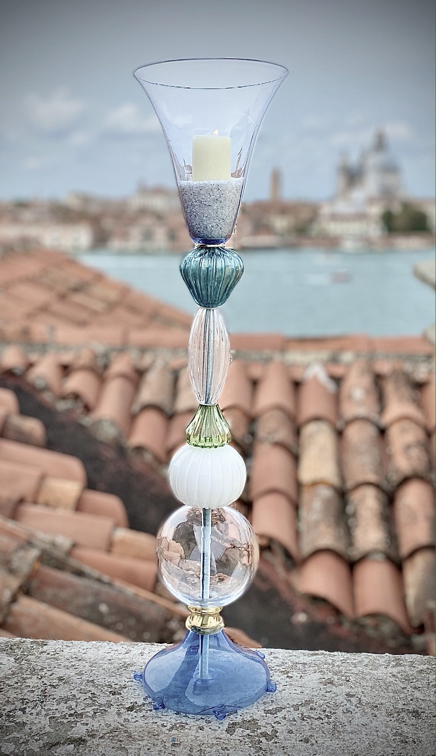 Tall glass candle holder with a white candle, decorative glass beads, and a river scene with historic buildings and rooftops in the background.