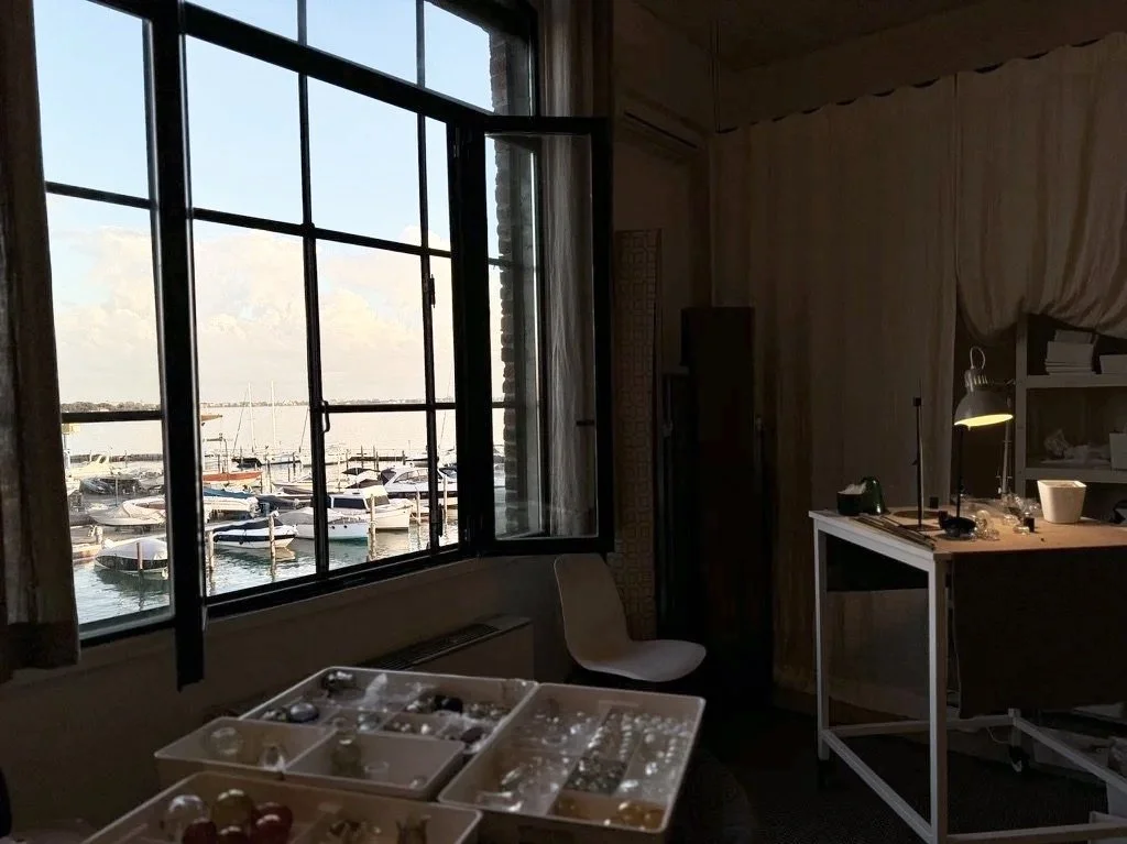 View of a marina with boats through a large open window in a dimly lit room with a white table, a white chair, and a desk lamp.