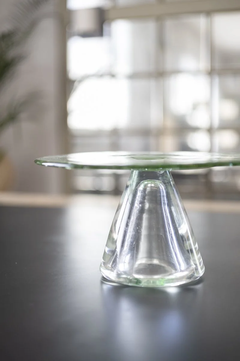 A clear glass plate balanced upside down on a glass cup, set on a black surface with a blurred window background.