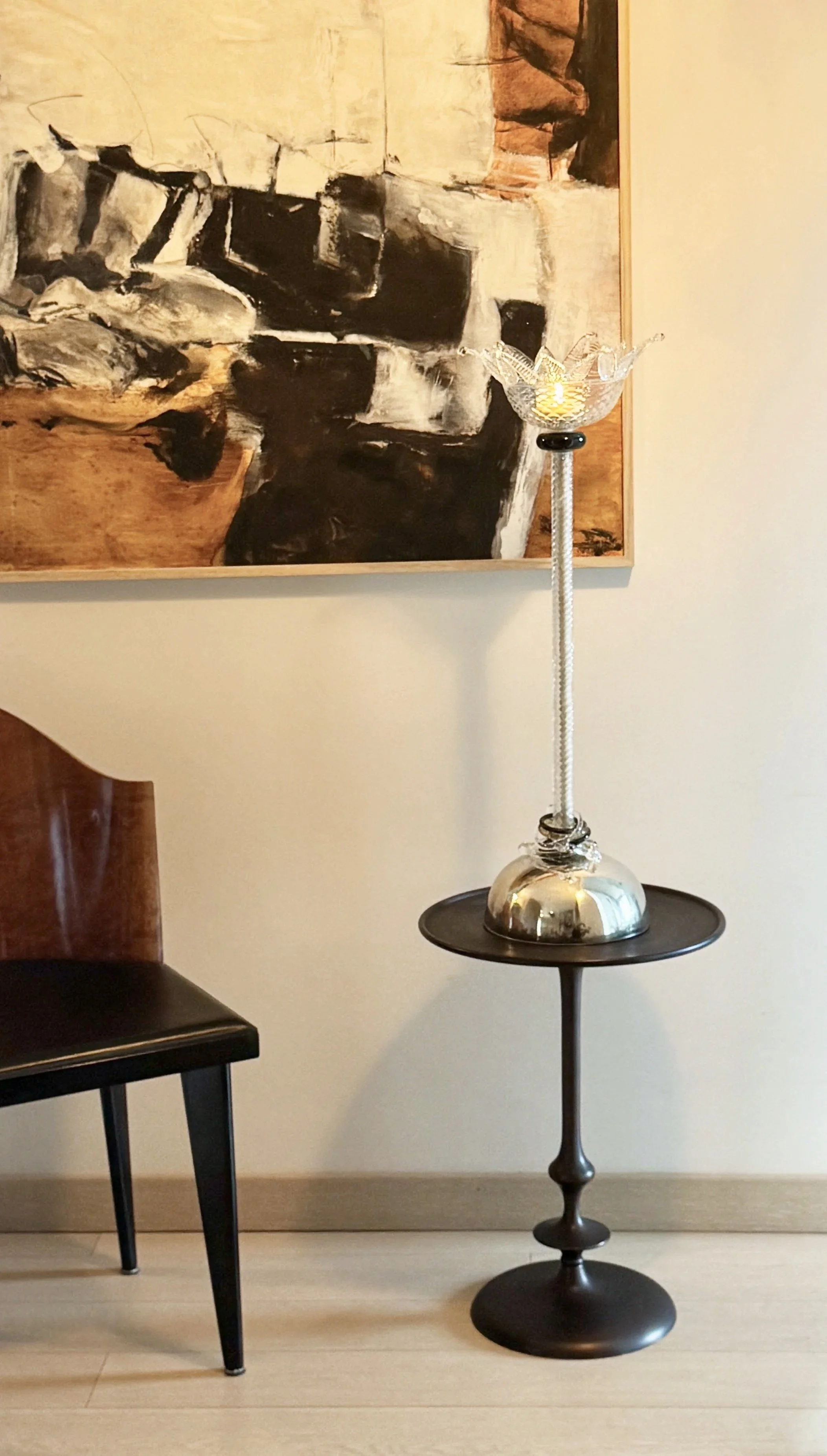 A decorative table lamp with a silver base and a crystal bowl-shaped top holding a candle, placed on a round black table. Behind is a framed abstract black and white painting on a light-colored wall, and part of a black chair to the left.