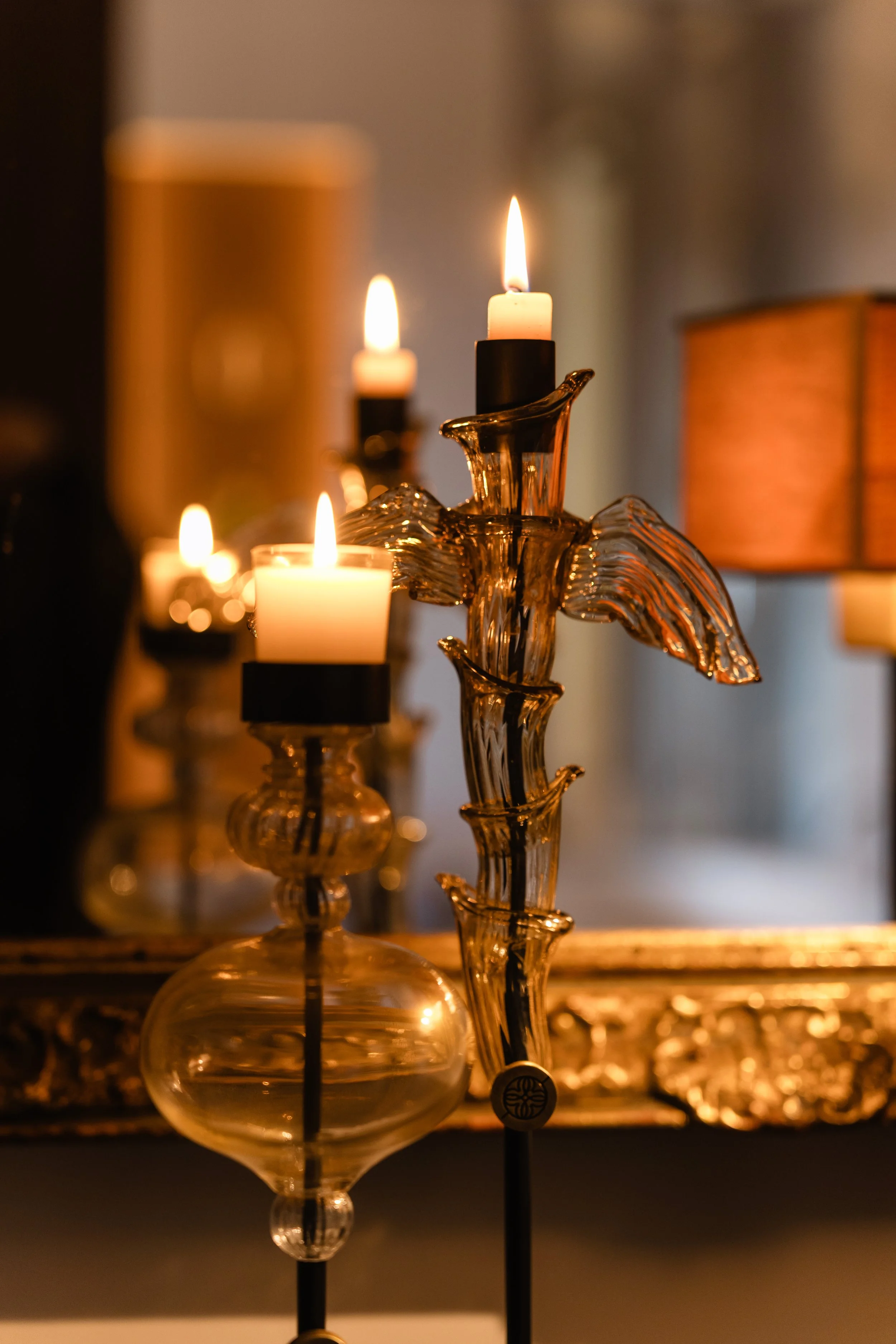Two glass candleholders with lit tapered candles, one with a decorative metal dove, on a reflective surface with a blurred warm-colored background.