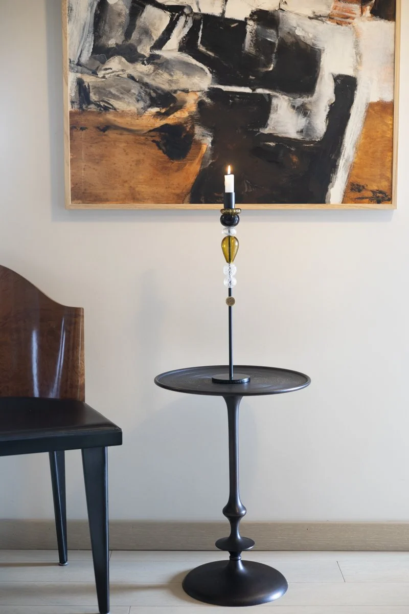 A modern black floor lamp with a candle holder design on a small round side table. In the background, there is an abstract painting with shades of black, white, gray, and brown on a beige wall. Part of a wooden chair with a black seat cushion is visi
