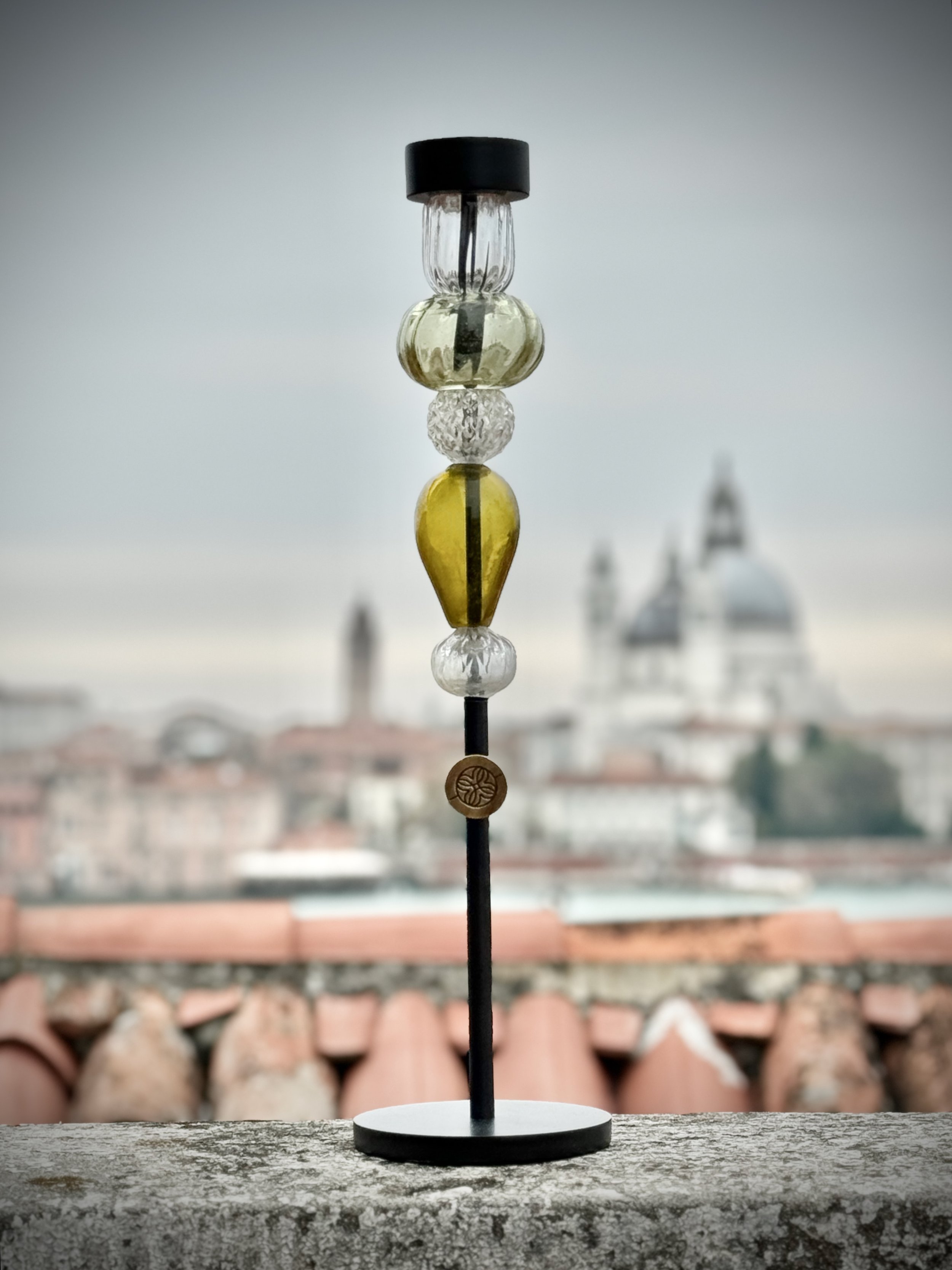 A decorative object with glass and metal beads on a black stand, placed on a weathered stone surface, with a blurred rooftops view in the background.