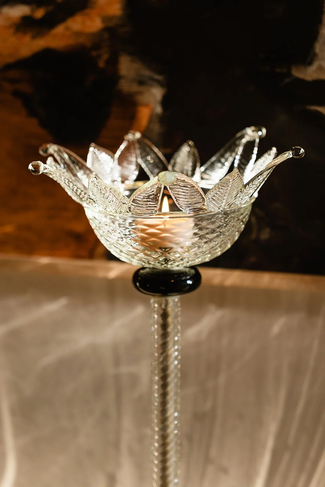 A decorative glass bowl with intricate patterns, holding a lit candle, is placed on a stand with a chain, against a dark background.