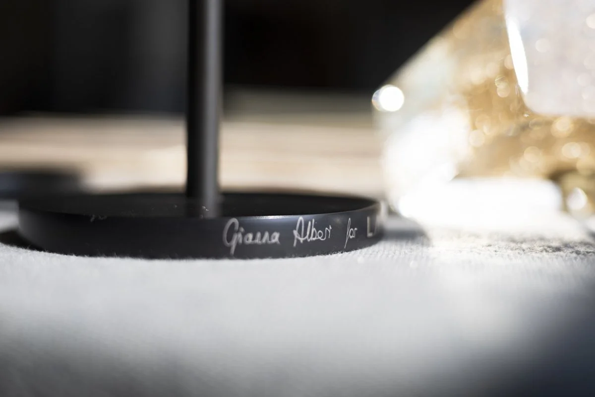Close-up of a black circular object with white handwritten text that says 'Giana Auction for Le' on a white surface, with a blurred glass object and other items in the background.