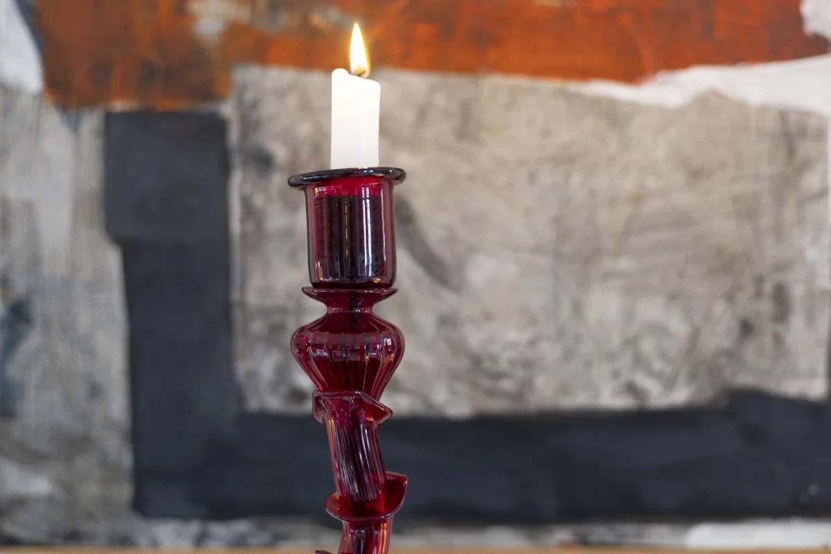 A lit white candle on a tall, red glass candle holder against a stone and wood background.