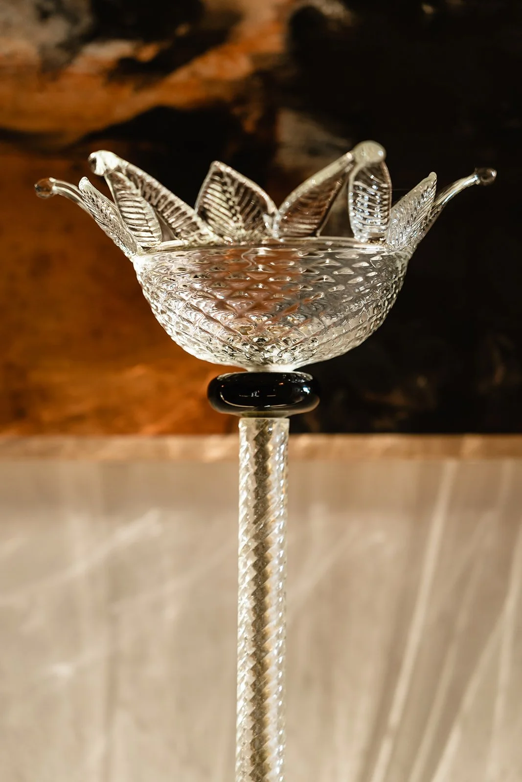 A clear glass sculpture resembling a bowl with leaf-like shapes, mounted on a textured glass stem against a dark background.