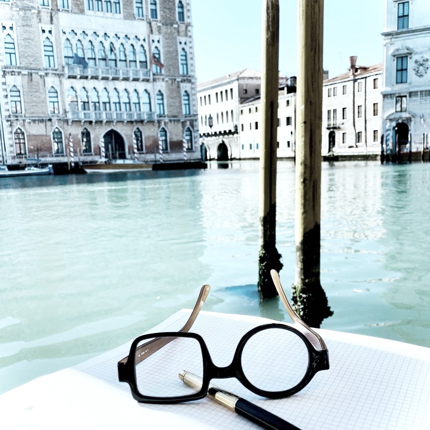 A pair of black eyeglasses and a pen resting on an open graph paper notebook, with a canal scene in Venice in the background, including old buildings with arched windows and wooden poles in the water.