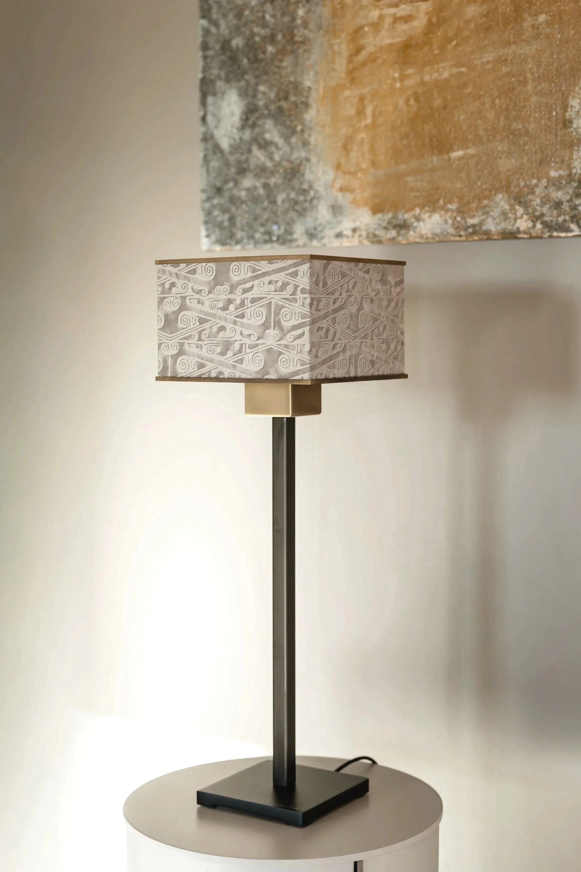 A modern table lamp with a rectangular base and a cylindrical stem. The lampshade has a geometric pattern with swirling lines in shades of gray and white.