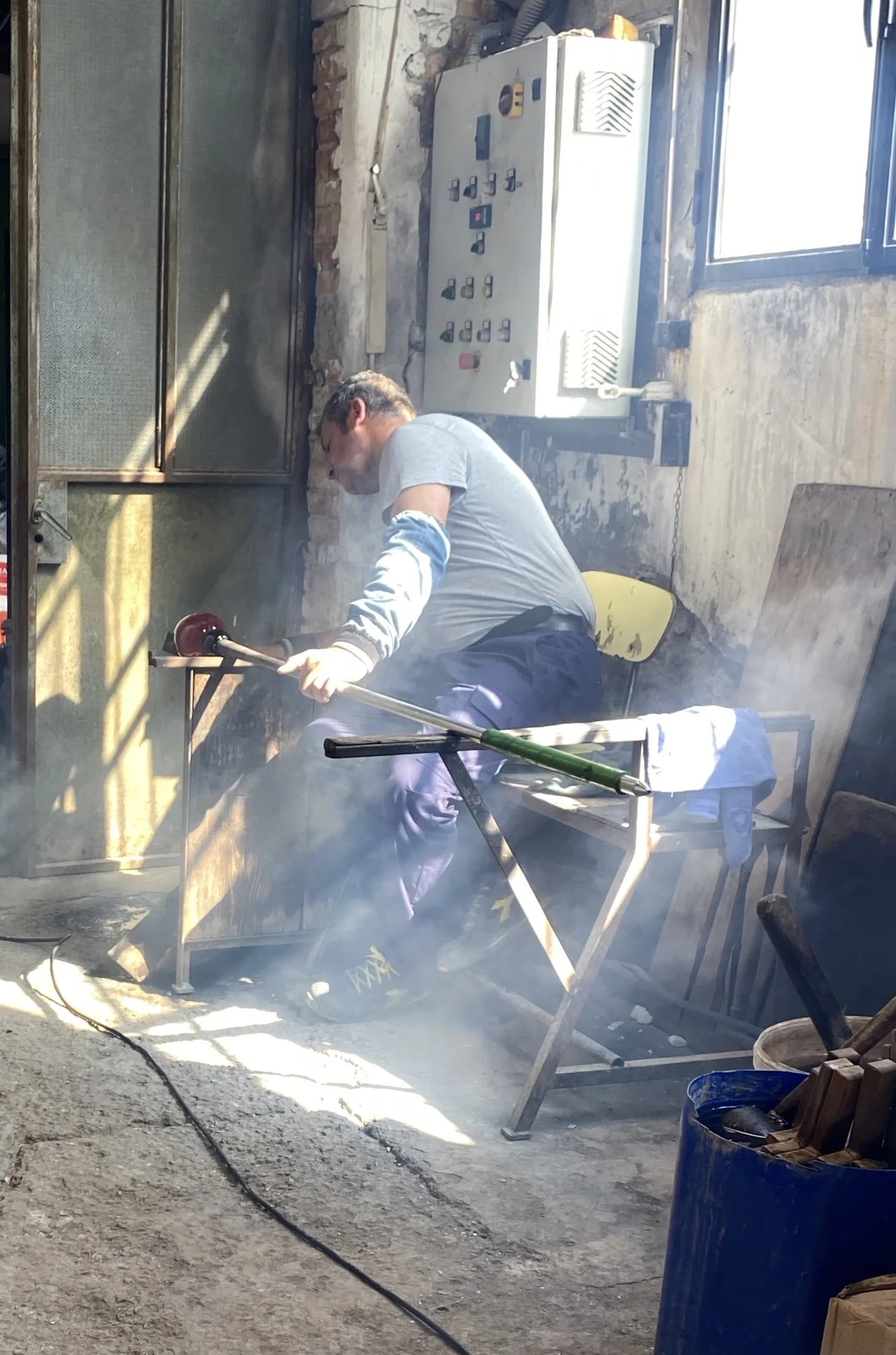 A man working on construction or renovation project, using a power tool with smoke or dust visible near his workspace. The setting appears to be an unfinished or partially renovated interior with exposed brick walls and various tools and equipment around.
