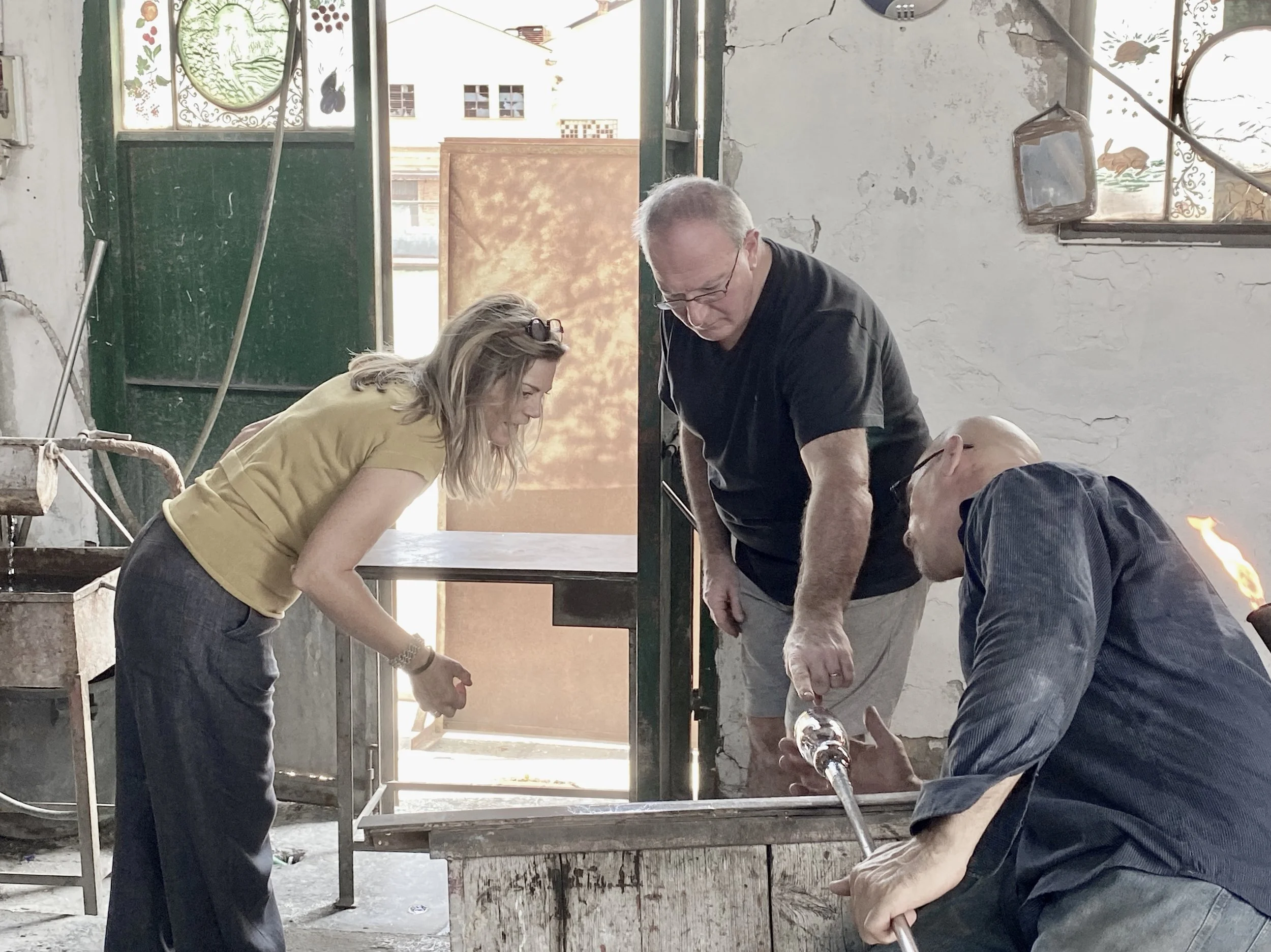 Four people, two women and two men, are working together in what appears to be a workshop or industrial setting, handling a glass object with a blowtorch. The space has a worn, rustic appearance with cracked white walls and a green door in the background, sunlight streaming through windows.