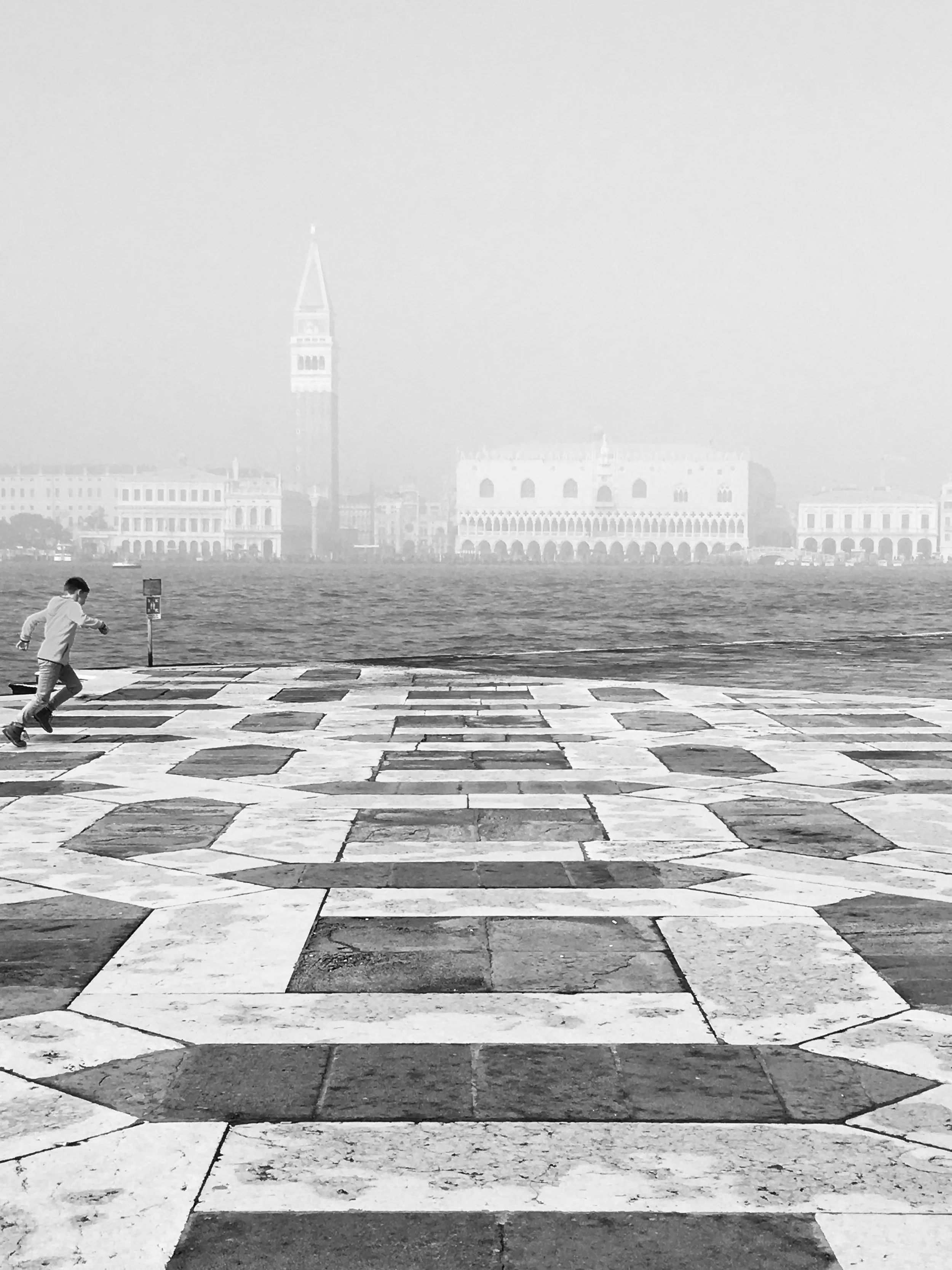 A child wearing a face mask is running across a stone-paved area near a body of water. In the background, there is a foggy view of a city skyline with historic buildings and a tall bell tower.