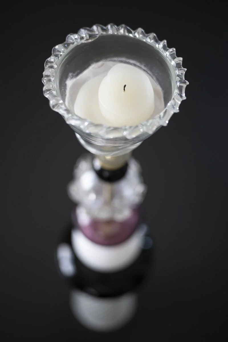 Close-up view of a white candle in a clear, decorative glass holder on a black surface, with the candle's wick slightly burnt.