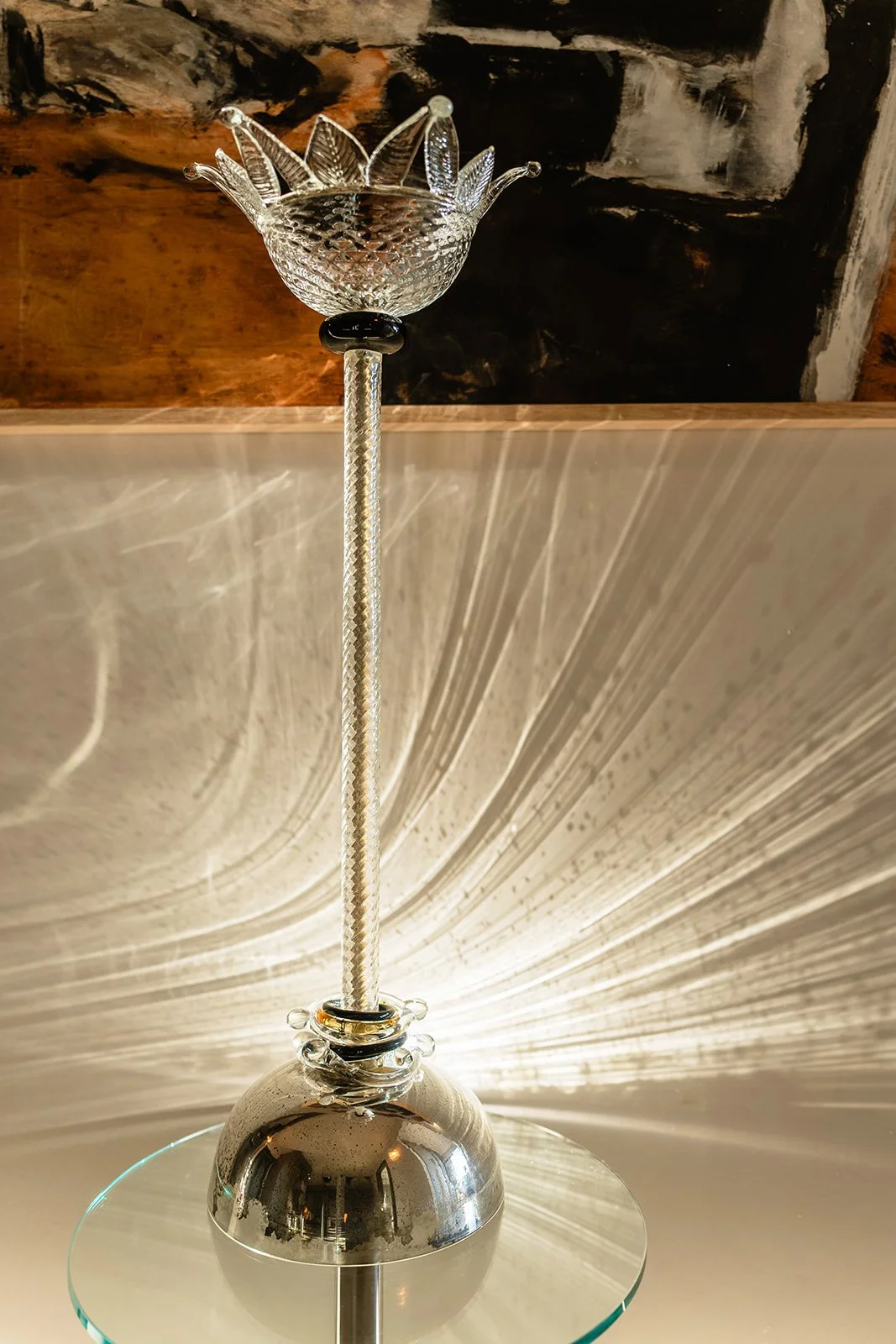A decorative glass sculpture resembling a crown or flower atop a tall, slender, textured metal stem, set on a circular mirrored base, with dramatic light reflections and a stone wall in the background.