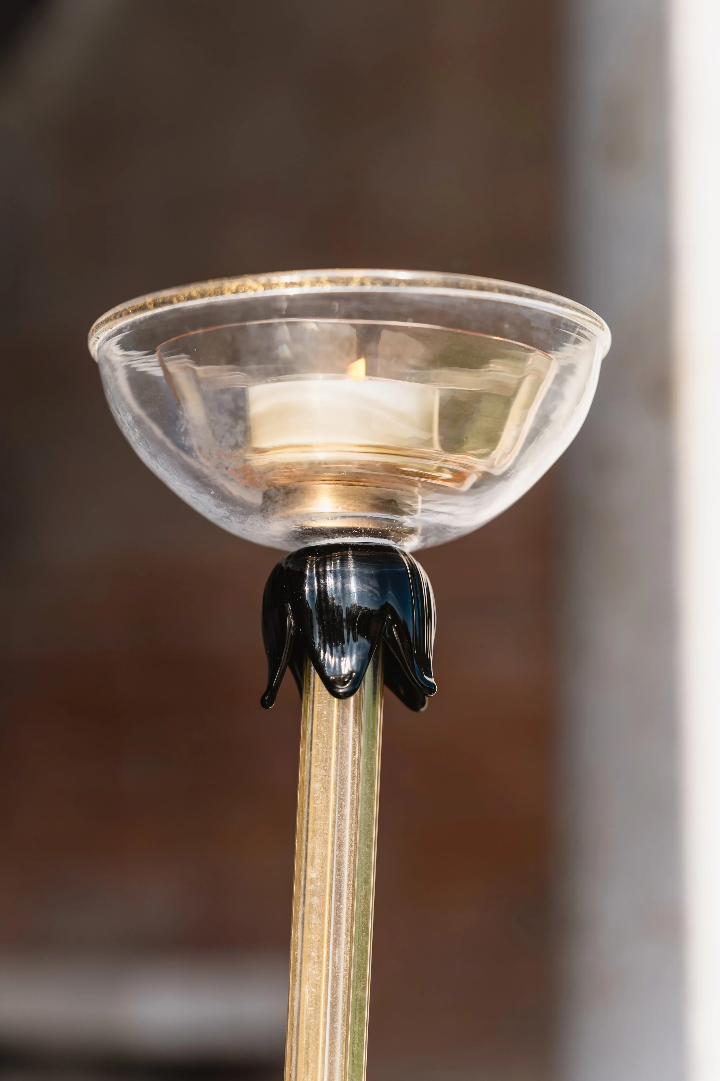 Close-up of a lit tealight candle in a glass holder with black floral decorations on a thin glass rod.