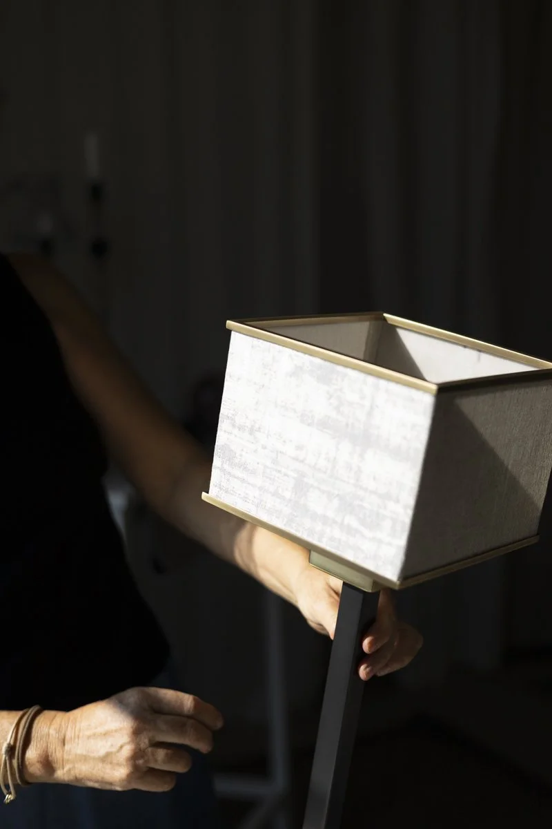 A hand holding a modern floor lamp with a rectangular fabric shade in a dimly lit room.