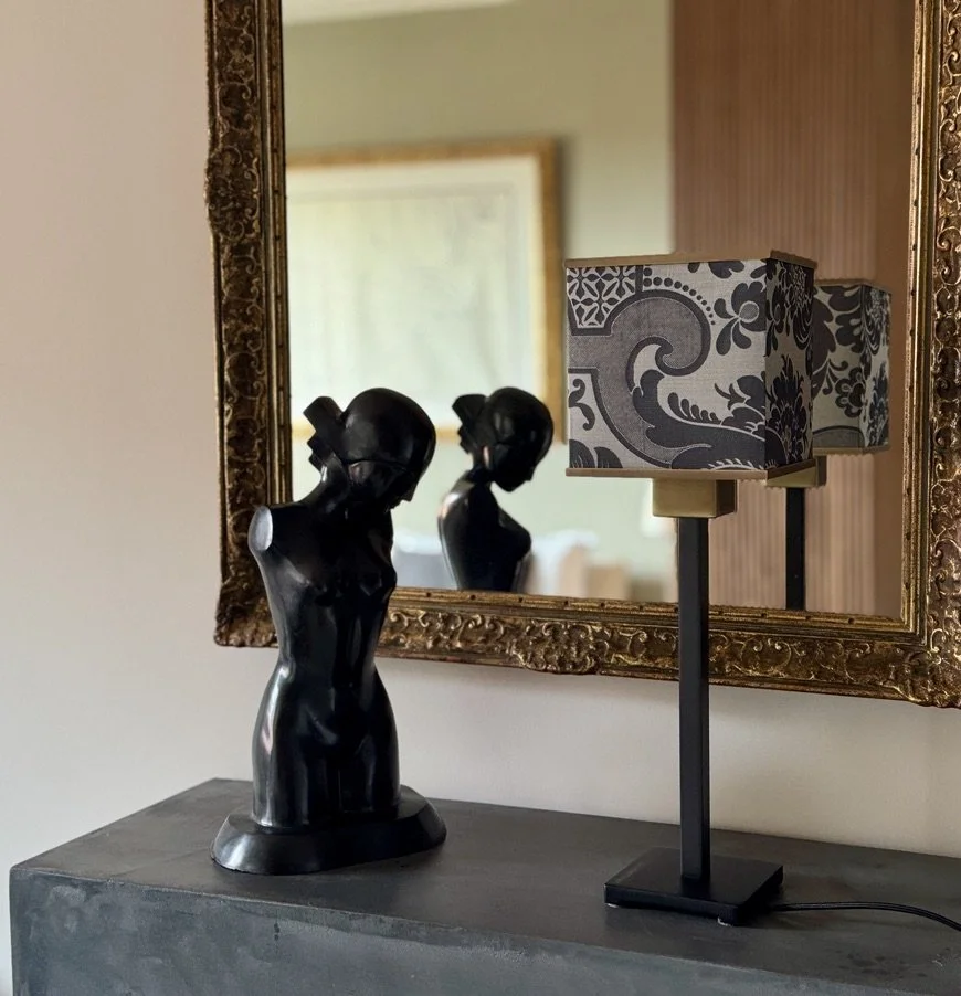 Decorative black bust sculpture of a woman's torso, modern table lamp with a patterned rectangular shade, and a mirror with an ornate gold frame reflecting the objects, all on a dark gray surface.