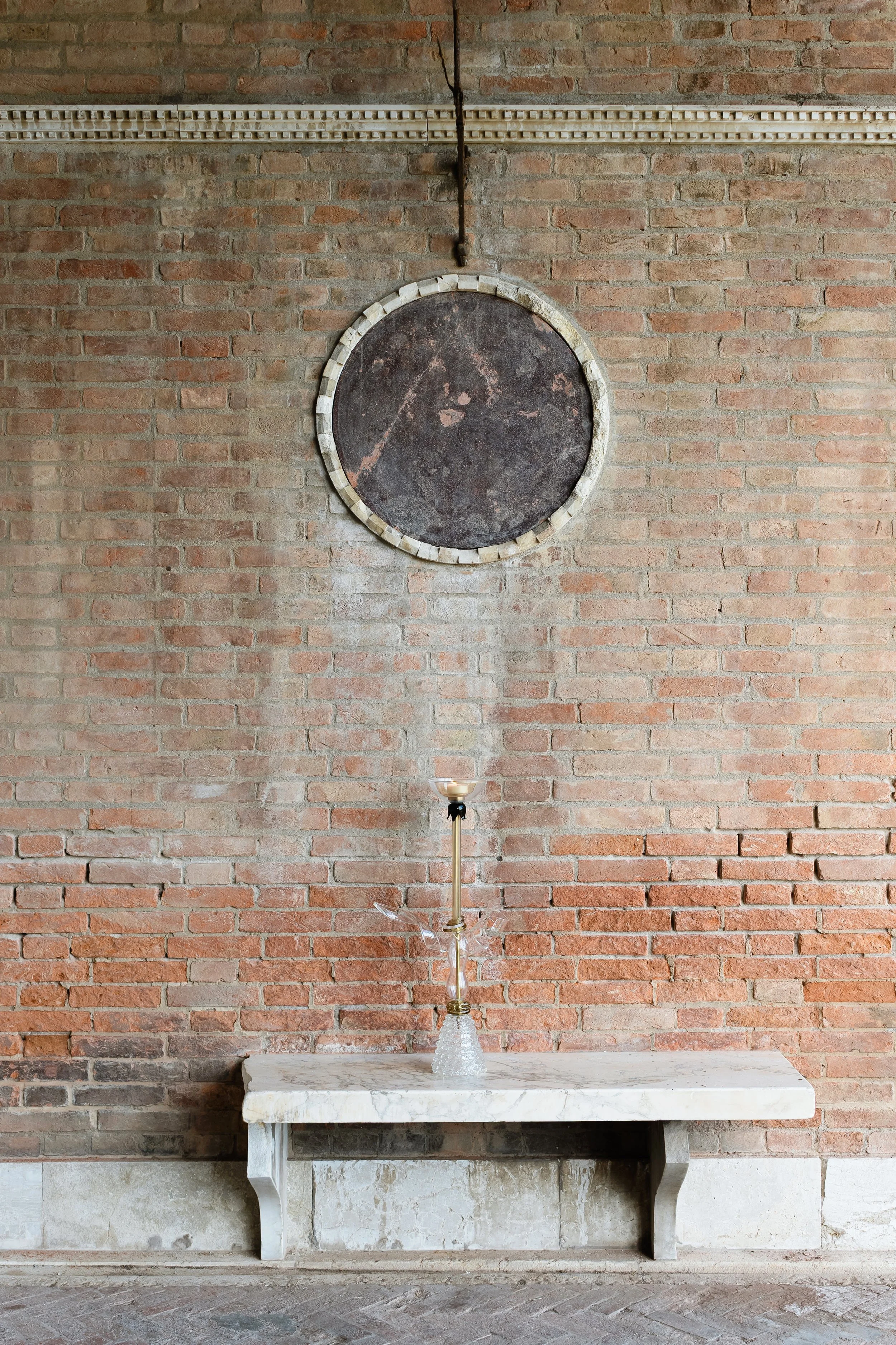A brick wall with a round black chalkboard and a white marble table with a glass chandelier in front.