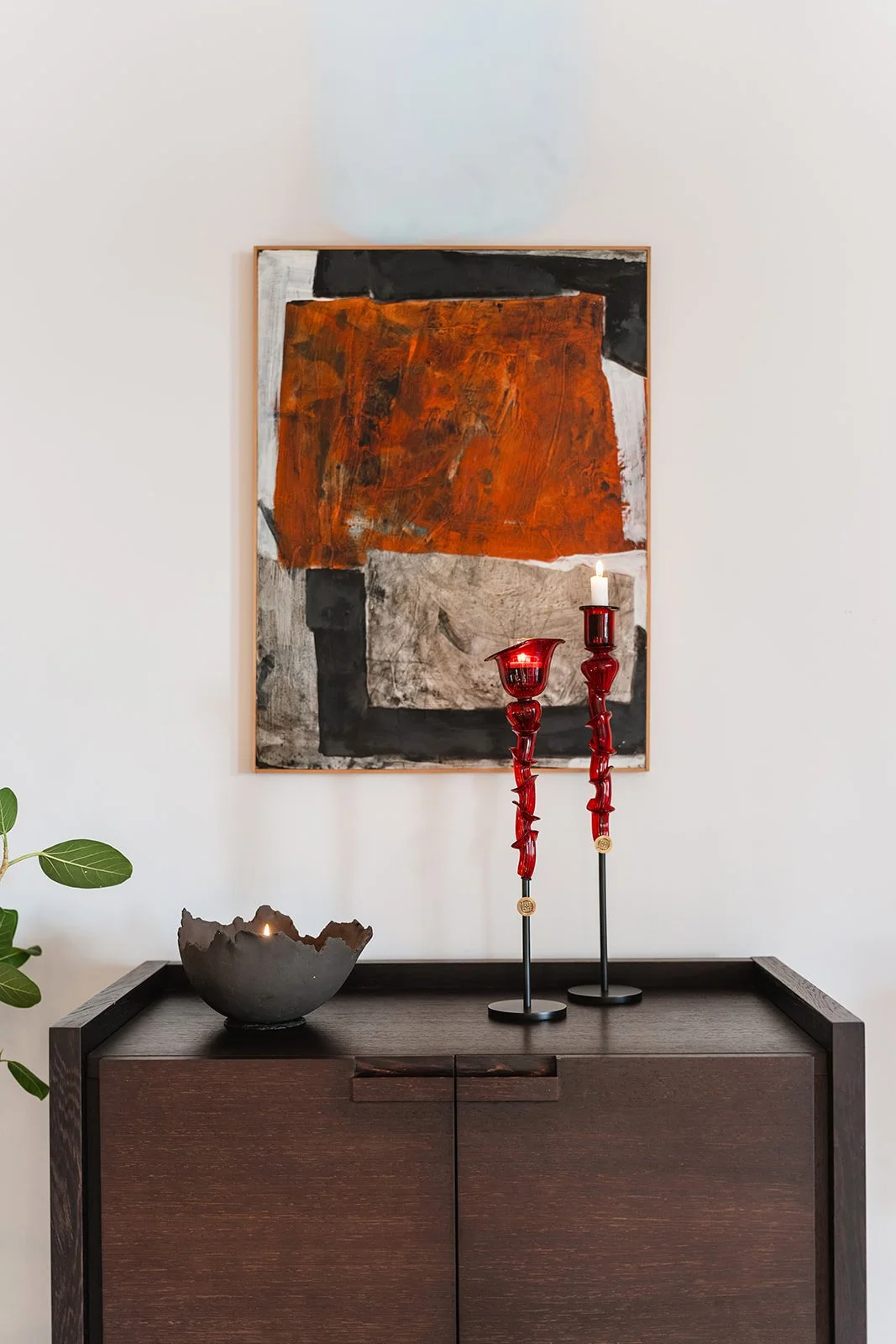Interior decor featuring a modern painting with bold black, orange, white, and gray abstract shapes hanging on a plain white wall. A dark wooden console table holds a decorative bowl with a lit candle and two red candle holders with candles. A green 