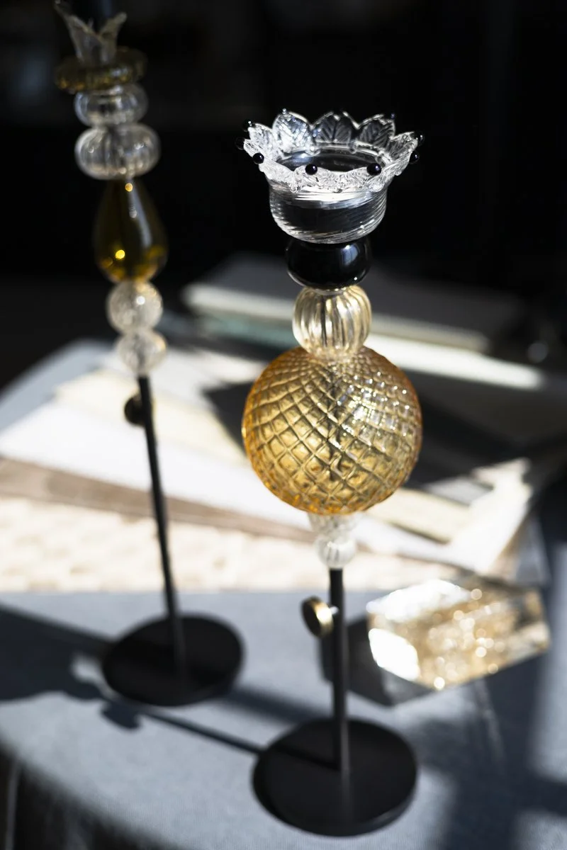 Decorative glass and metal stands with ornate glass tops and spherical glass bodies, displayed on a table with a cloth and papers.