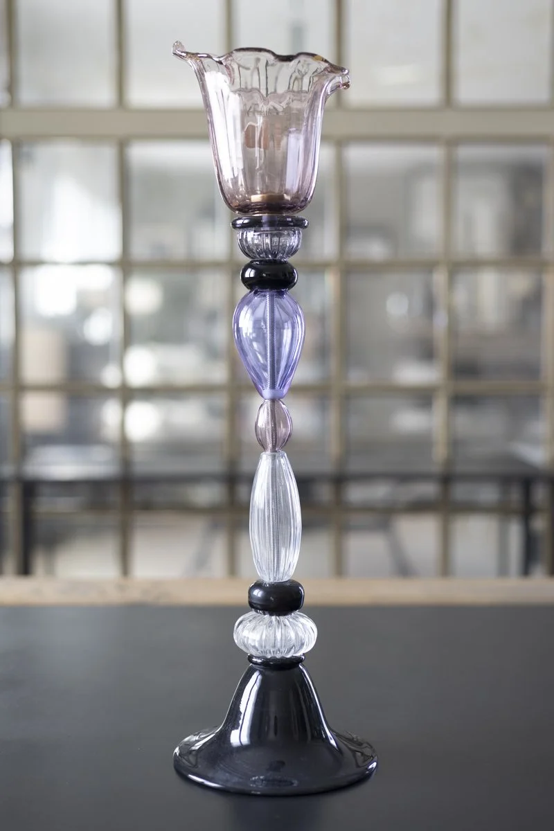 A tall glass candle holder with a pink, purple, clear, and black color scheme, featuring various shaped glass segments stacked vertically, with a flared top.