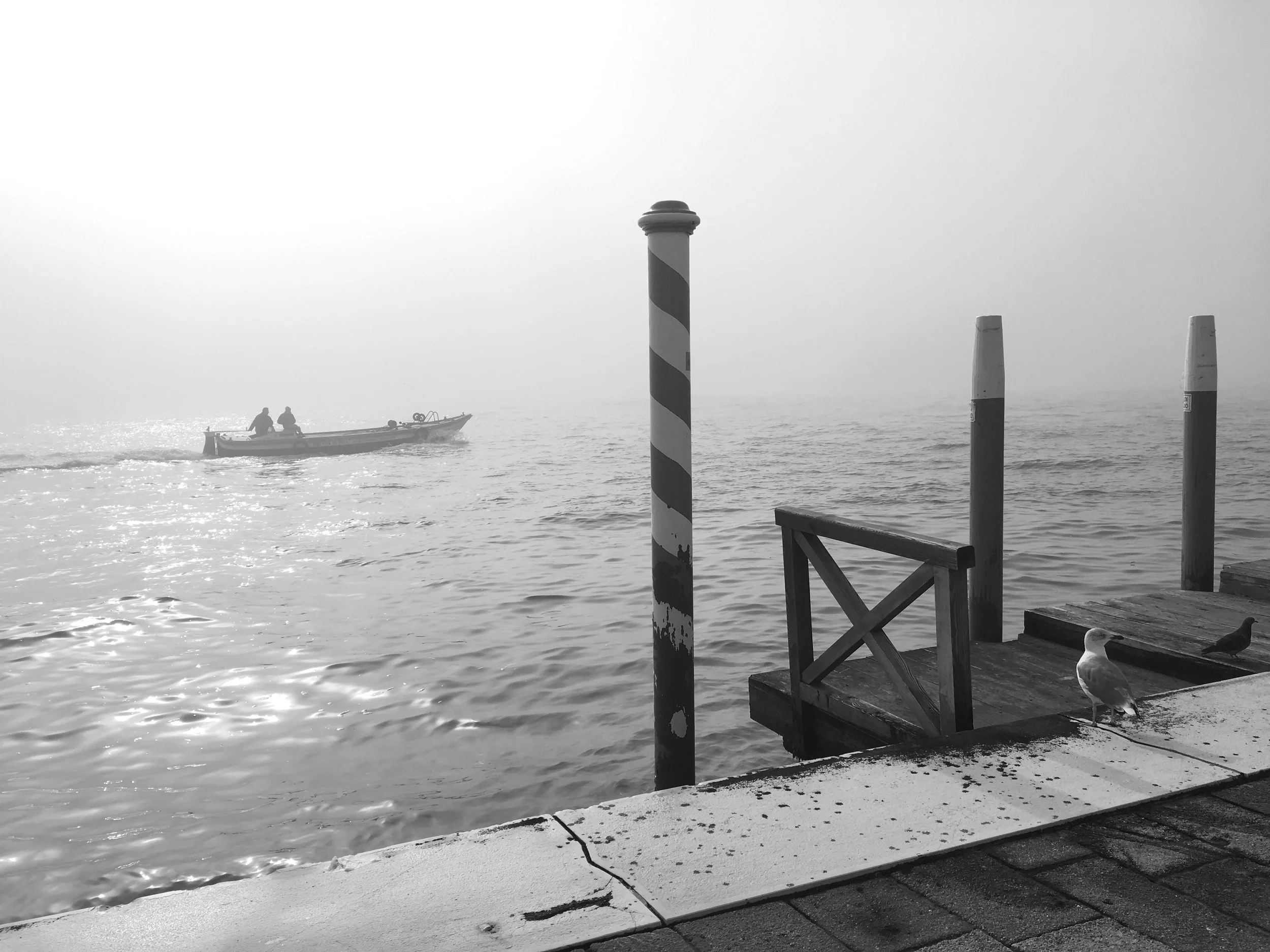 A black and white photo of a waterfront scene with a boat carrying two people, a seagull standing on the dock, and two thick striped mooring posts near the water.