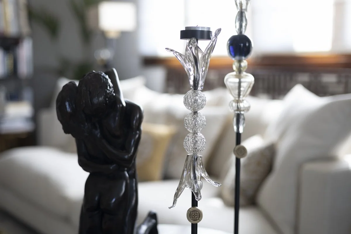 Close-up of two ornate decorative objects in a living room, including a black sculpture and a glass sculpture with three large crystal-like spheres, with a blurred beige sofa and window in the background.