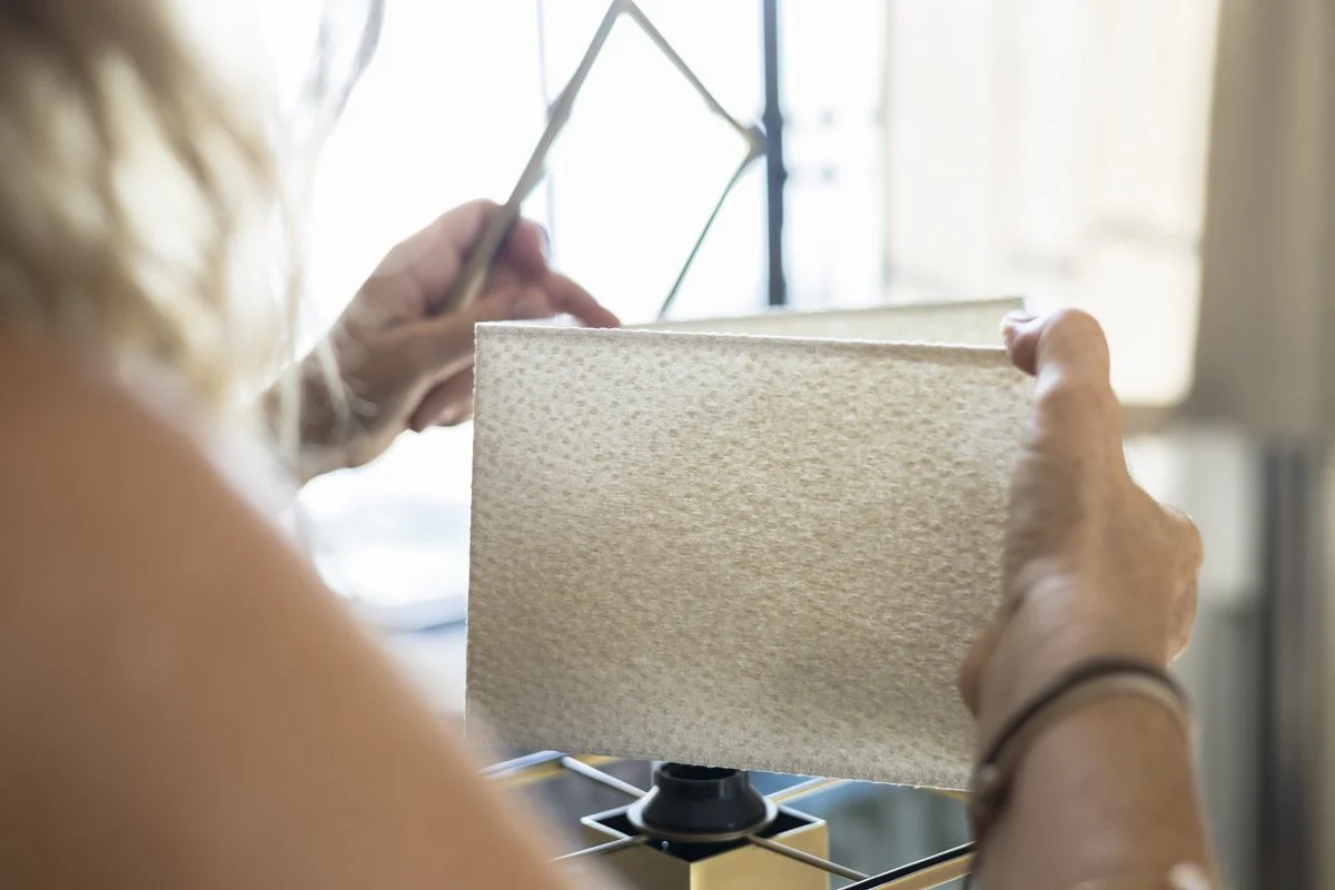 A person holding a piece of textured fabric while working on a lamp with a rectangular shade in a well-lit room.