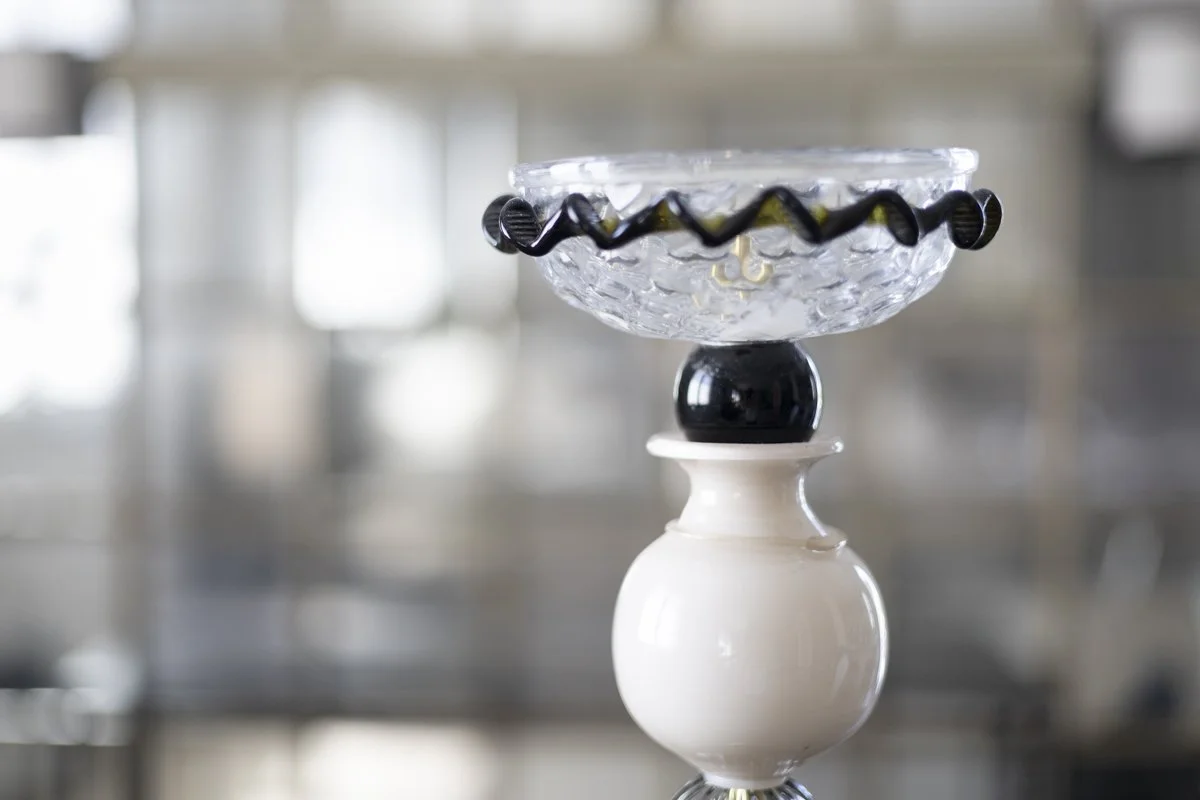 Glass bowl with ice cubes and black and yellow decorative ribbon, balanced on a black and white ceramic pedestal, in a blurred background.