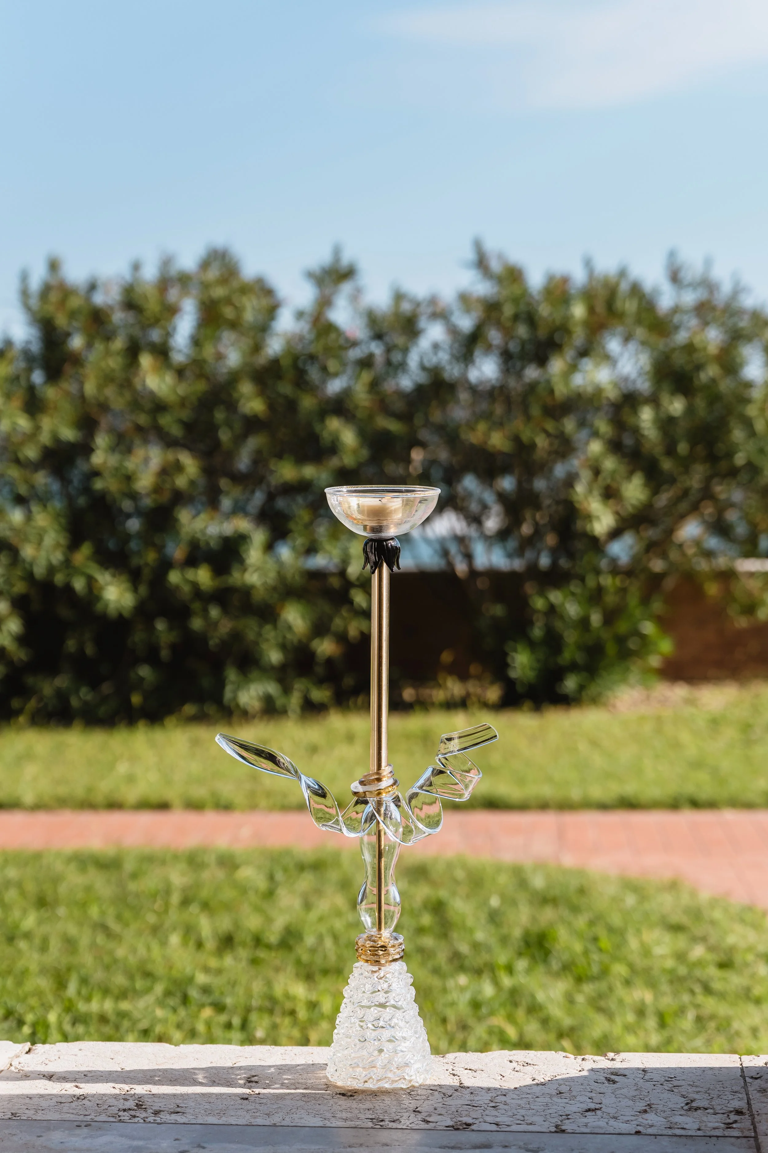 Glass sculpture resembling a bird or dragonfly with a long neck and decorative wings, placed outdoors on a concrete surface with a background of trees and blue sky.