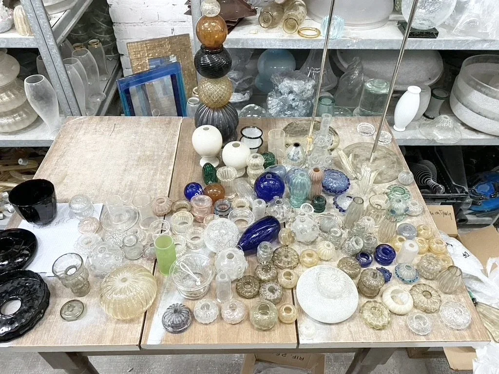 Table filled with various glass and ceramic objects, including vases, bowls, and ornaments, in a cluttered store setting.