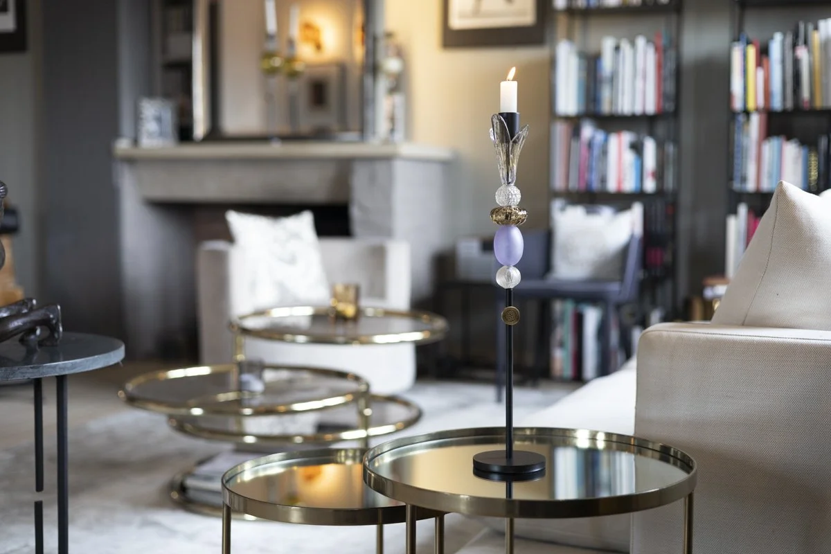 Living room with a gold-trimmed round side table, a candle holder with a lit candle, a bookshelf filled with books, and comfortable seating including a white sofa and a white armchair.
