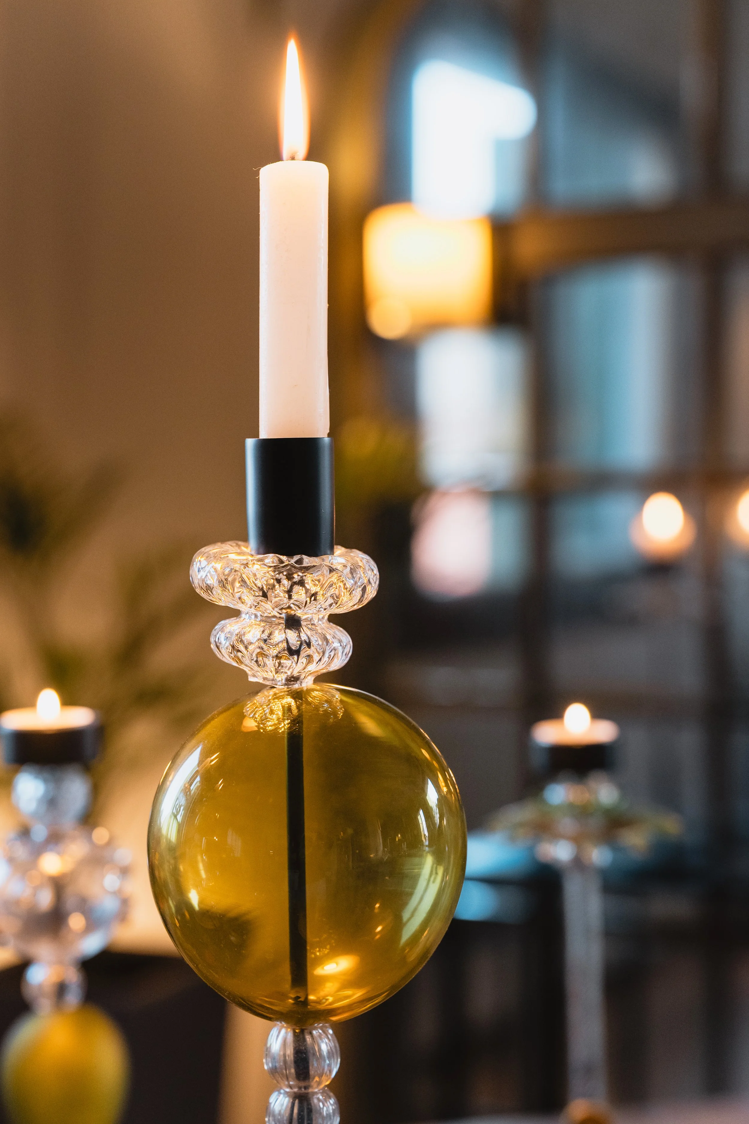 Close-up of a lit candle in a black holder, set on top of a decorative glass ornament that is yellow in color, with blurred background of additional candles and window.