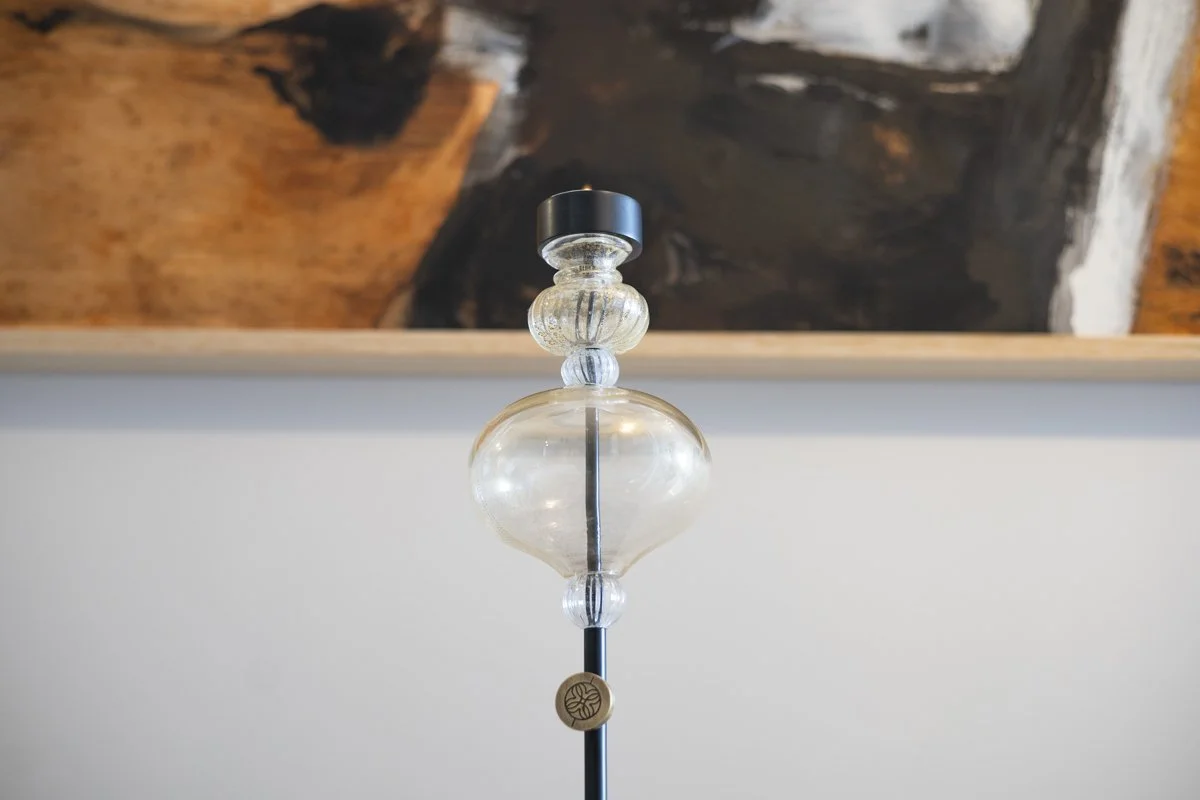 Close-up of a glass lamp with a wooden flower-shaped adornment on the black metal pole, set against a white wall with an abstract wooden art piece in the background.