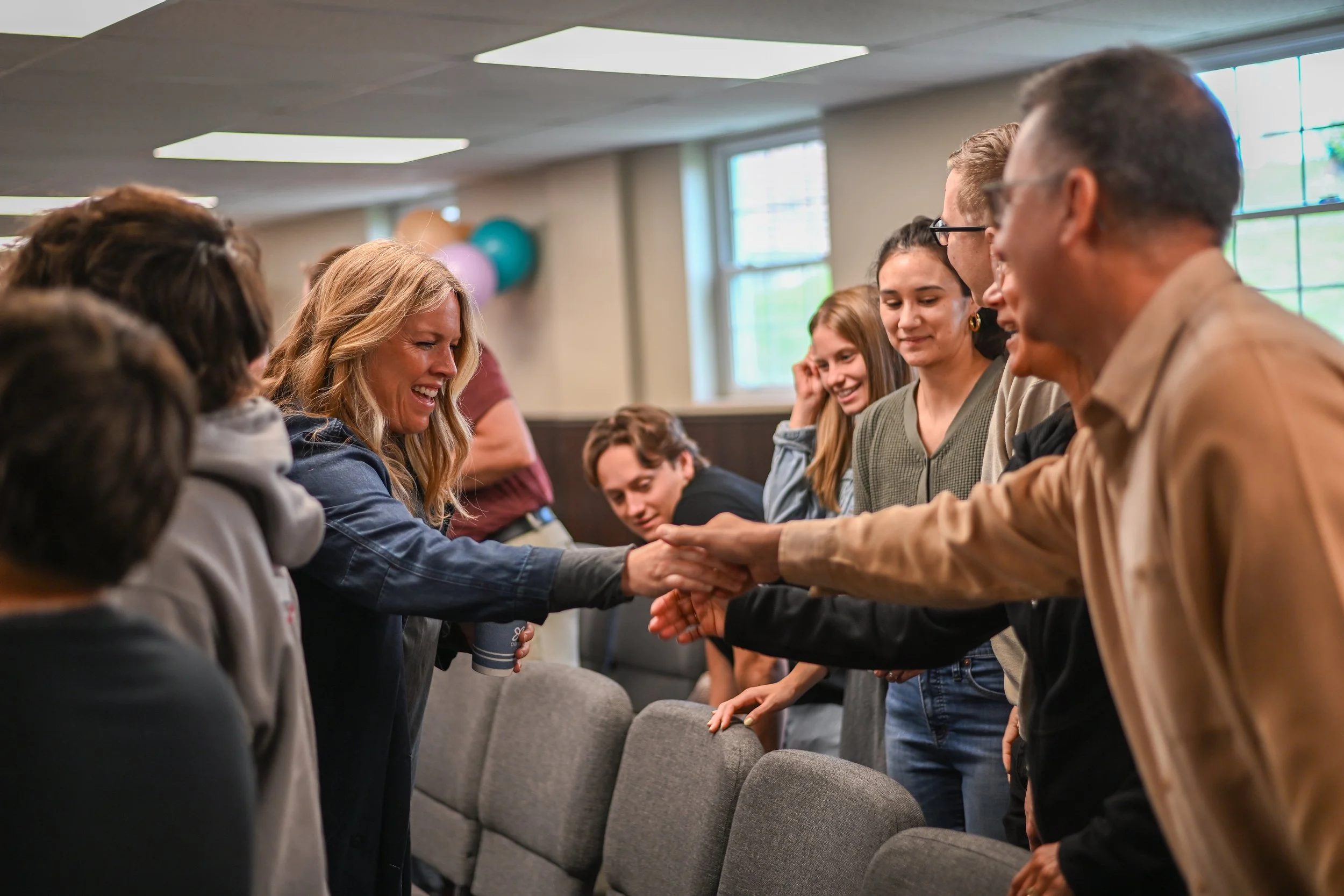 Group of people standing in a room, engaging in a handshake and conversation; some are smiling, and balloons are visible in the background.