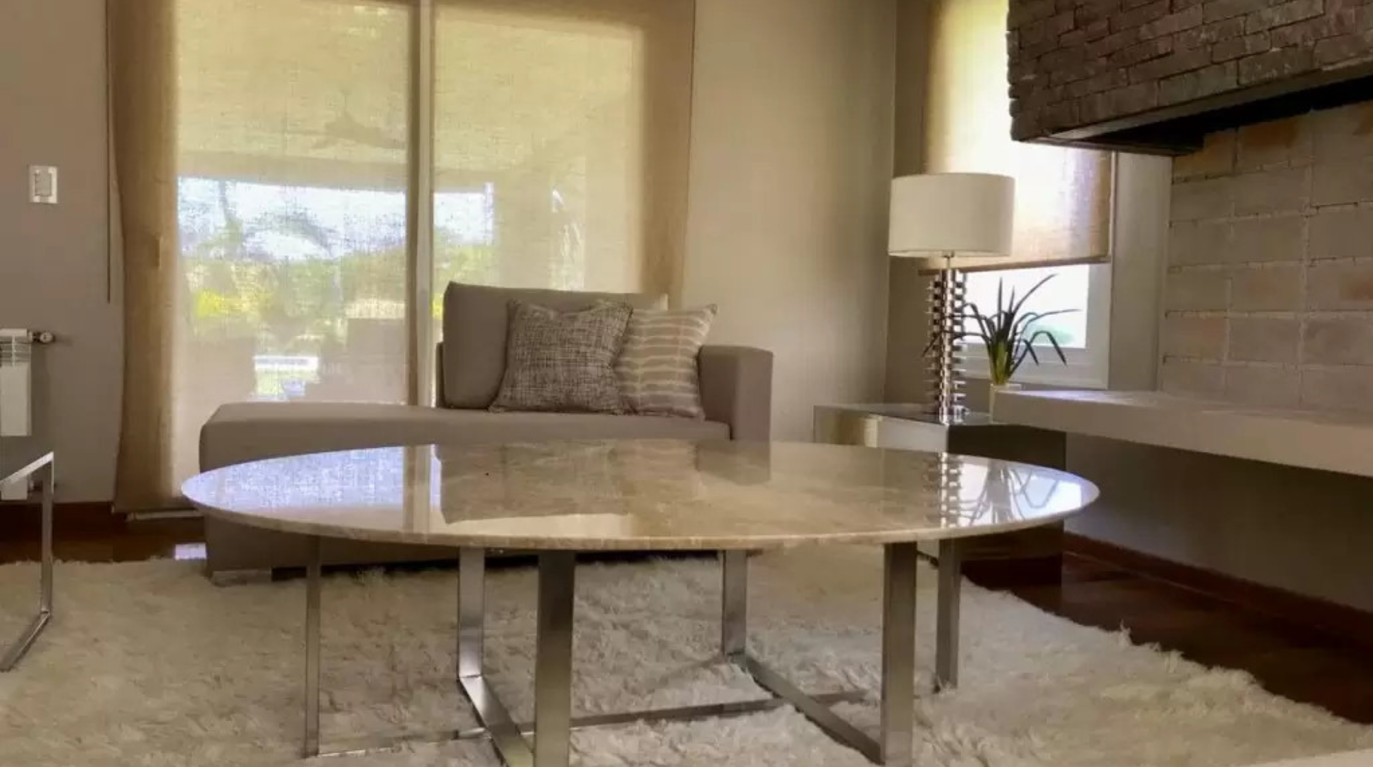 Sala de estar con sillón, mesa de centro de mármol, lámpara y plantas, con cortinas beige y muro de ladrillo visto